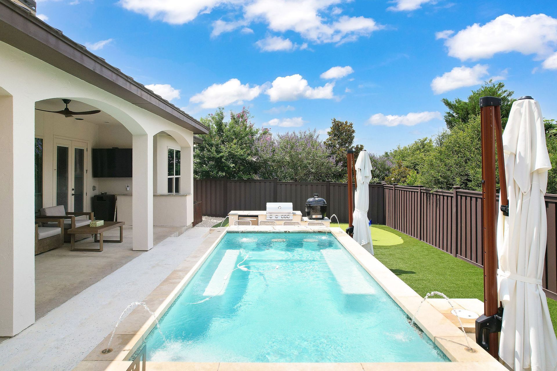 Backyard pool beside a white house, with lounge chairs, fence, and blue sky with clouds.