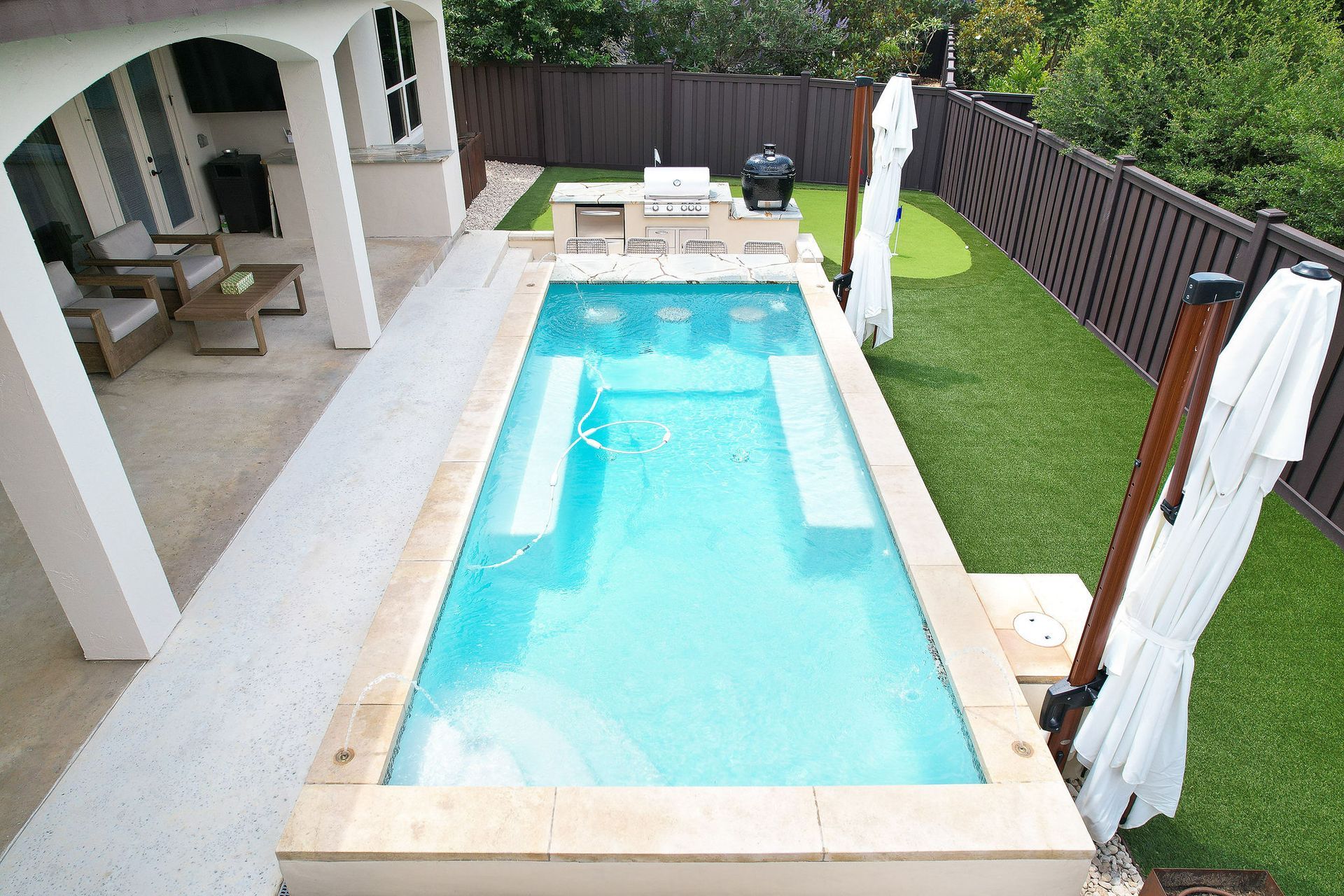 Narrow turquoise backyard pool with lounge chairs, patio, and green turf beside a house