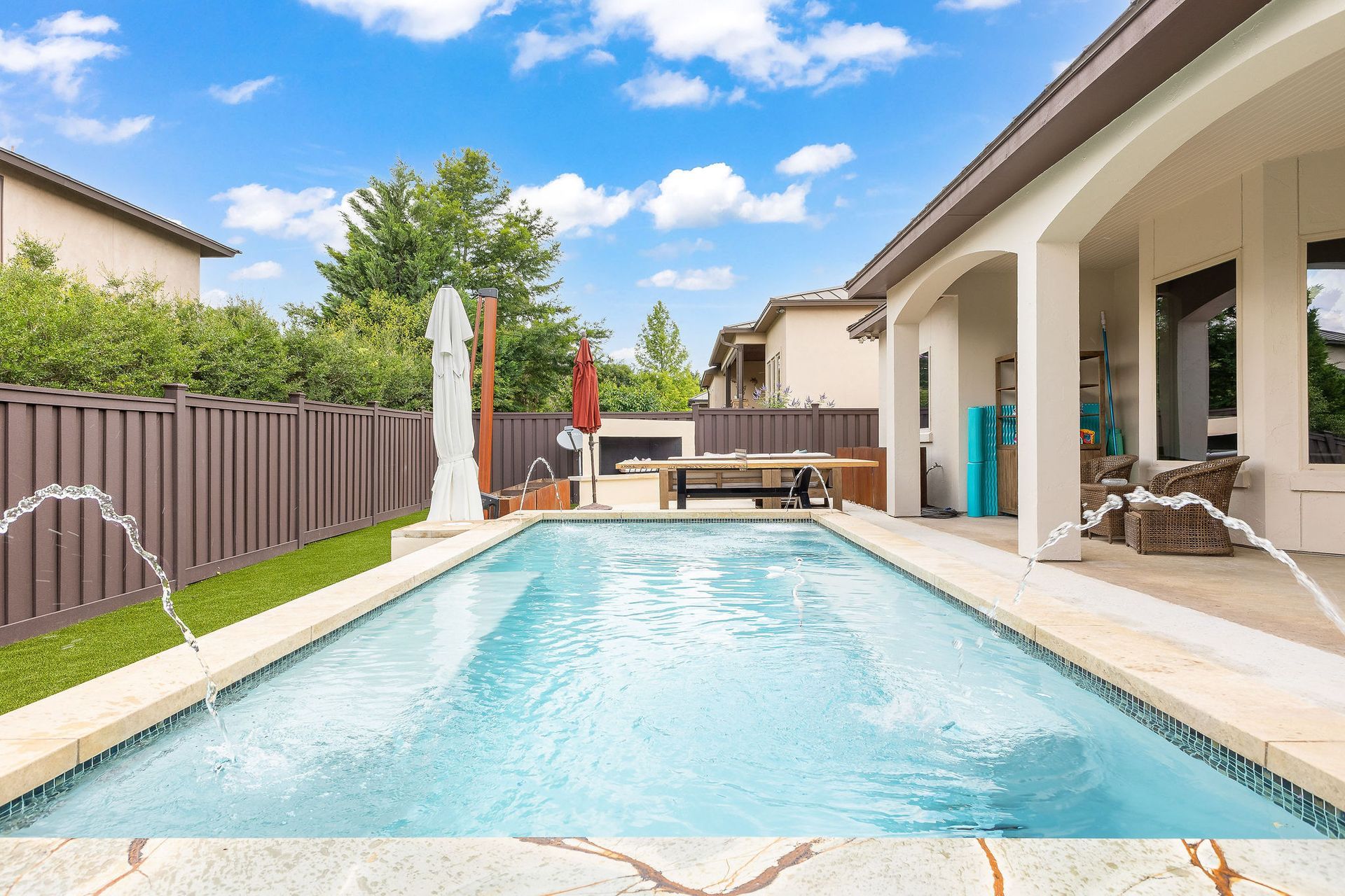 Backyard pool beside a covered patio and fenced lawn under a blue sky