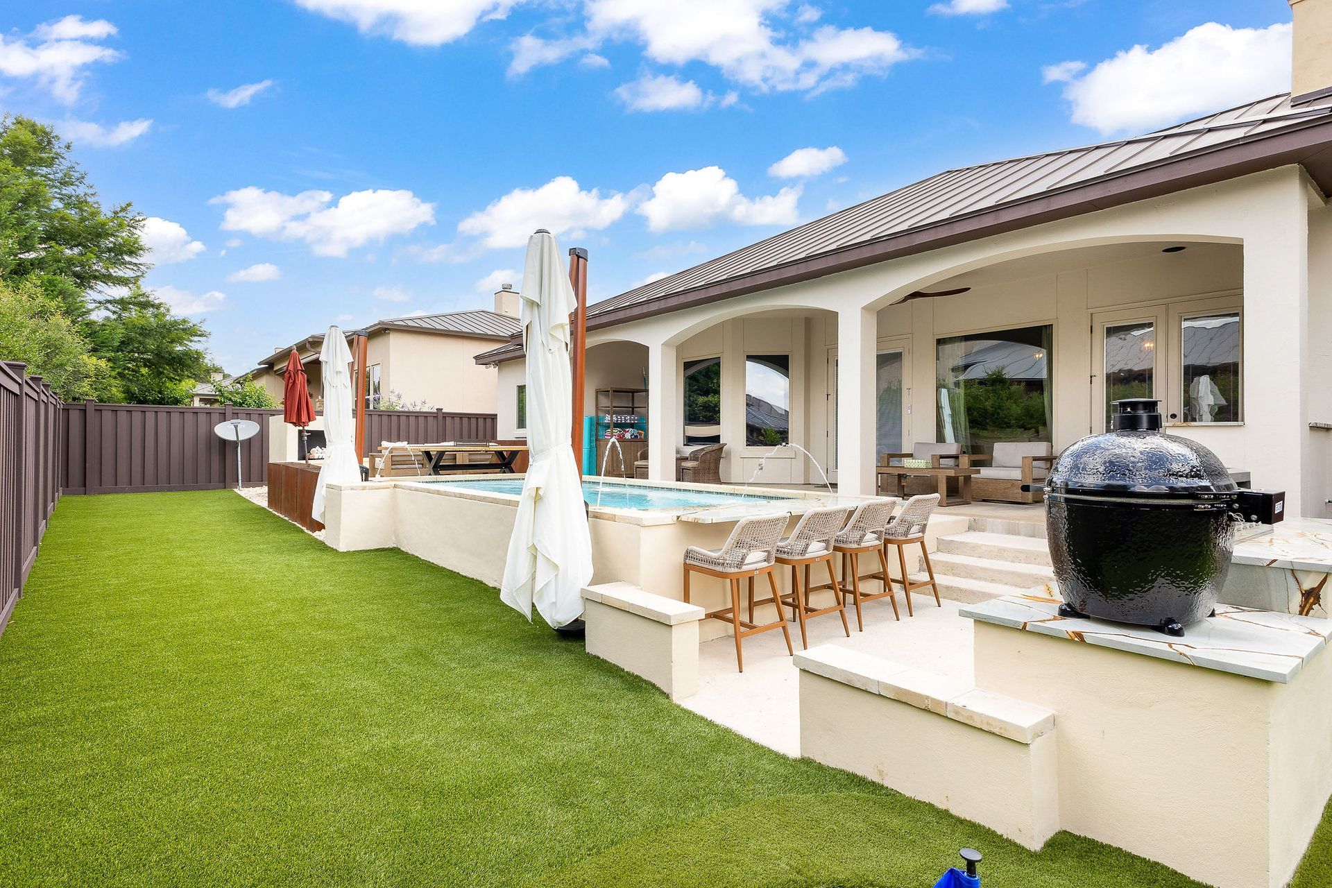 Backyard patio with green lawn, covered seating area, and a black barbecue grill under a blue sky