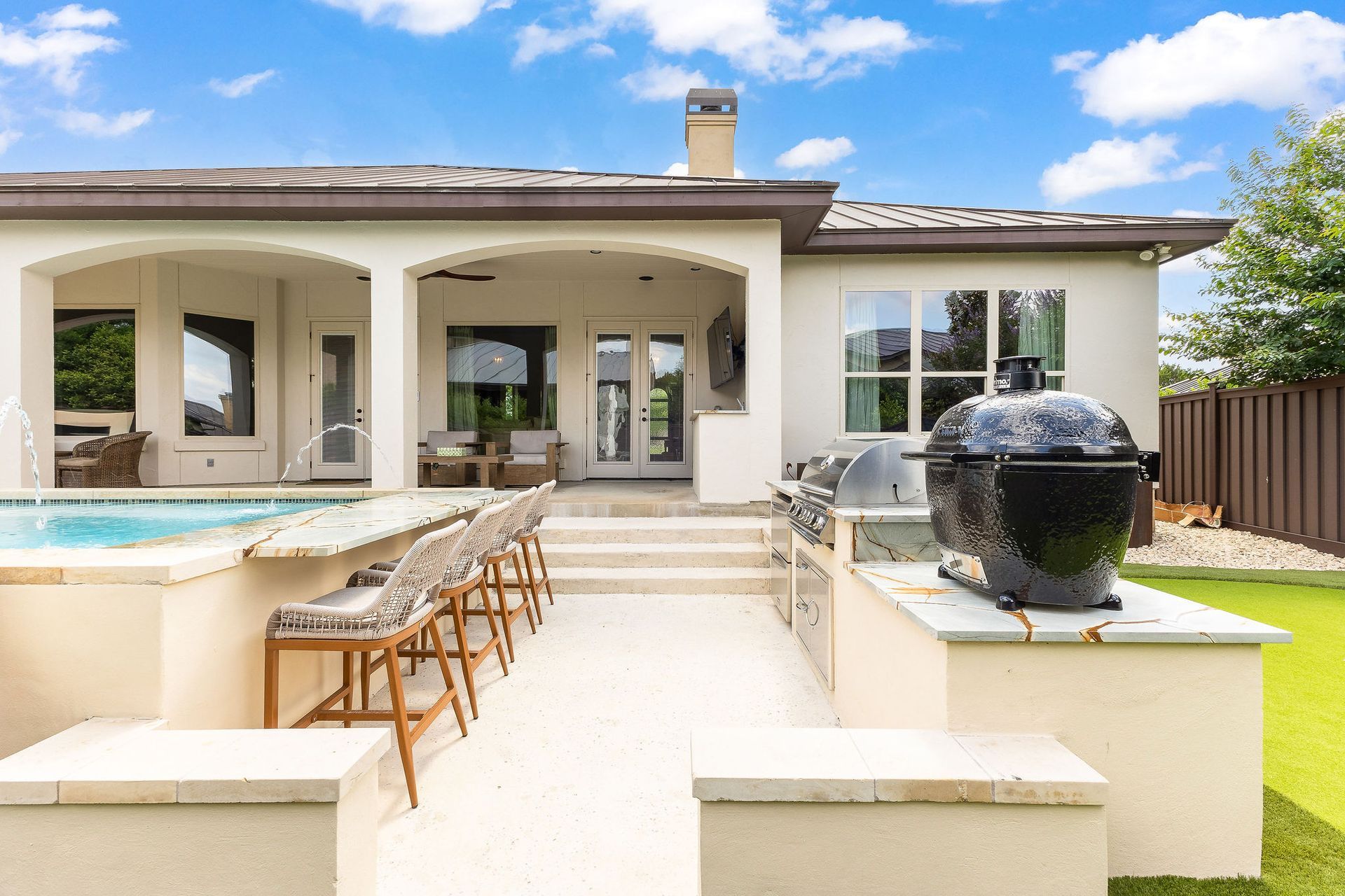 Modern backyard patio with pool, outdoor dining table, and black grill beside a cream-colored house