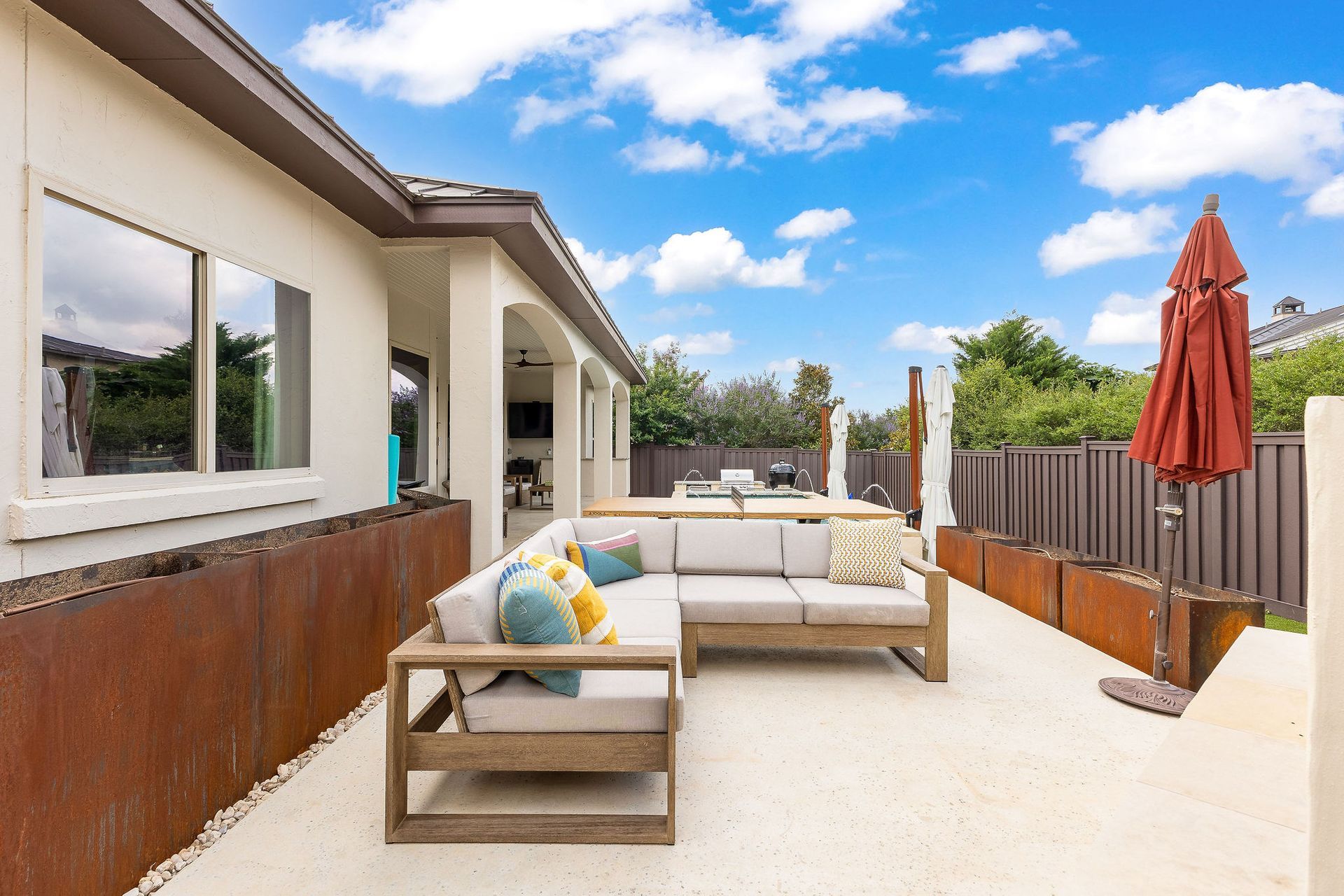 Patio with beige outdoor sofa, wooden fence, and red umbrella beside a house under a blue sky