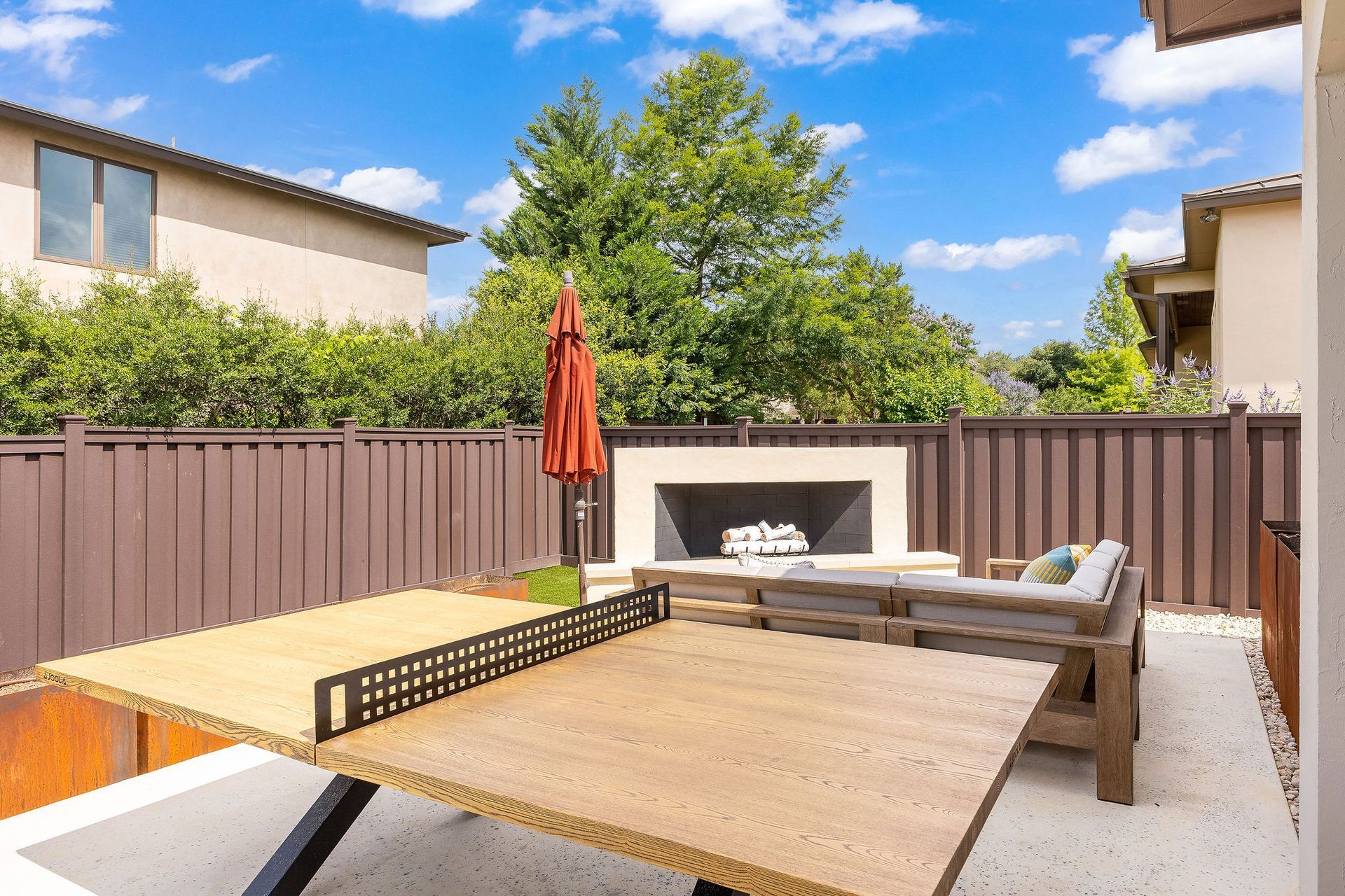 Backyard patio with wood fence, outdoor dining table, lounge chairs, and fire pit under blue sky