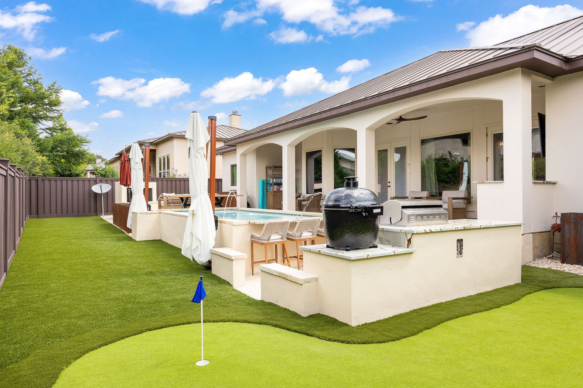 Modern backyard patio with white stucco house, covered seating, and green lawn under a blue sky
