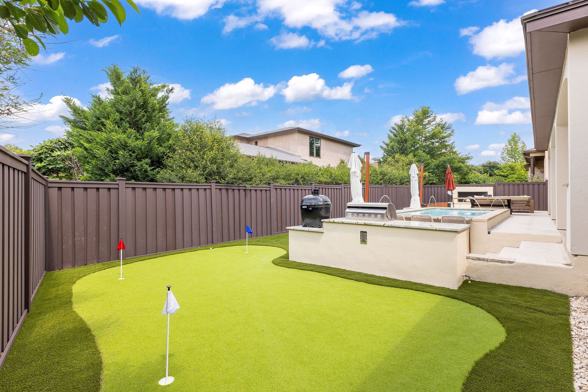 Small backyard putting green with a flag, patio seating, and a wooden fence under a blue sky.