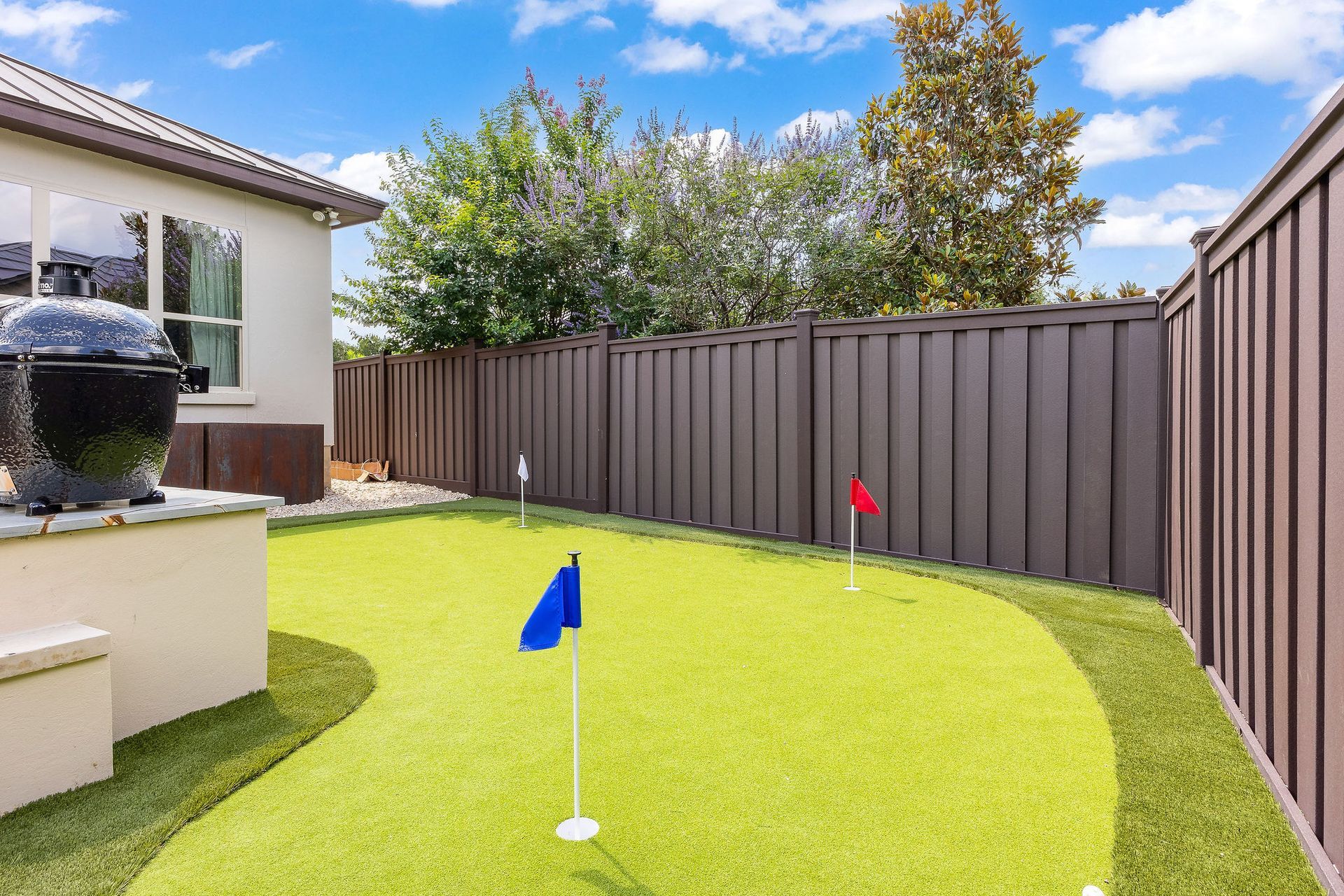 Small backyard with artificial grass, brown fence, and two mini golf flags under a blue sky.