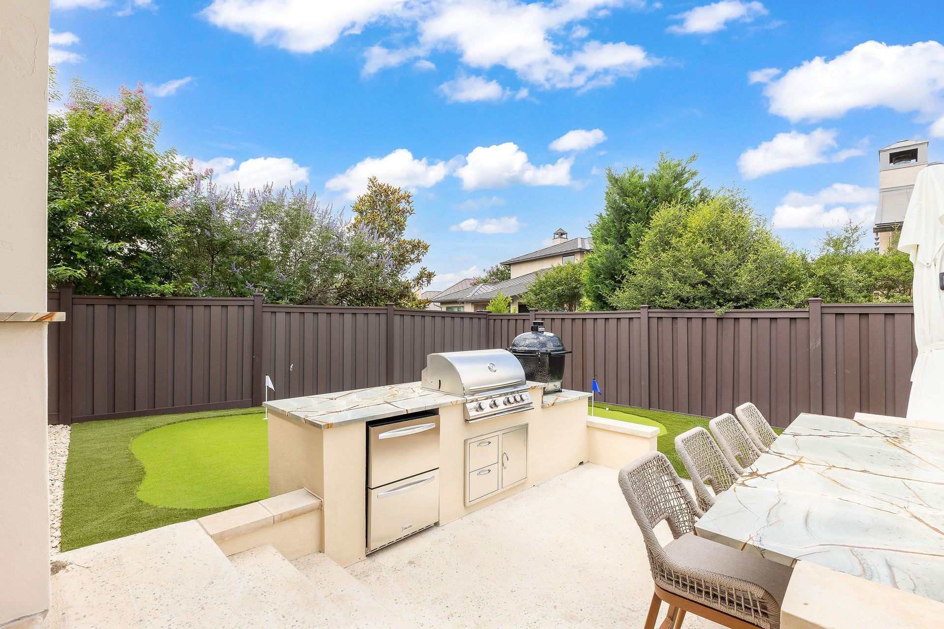 Outdoor patio with grill island, dining table, and fenced backyard under a bright blue sky
