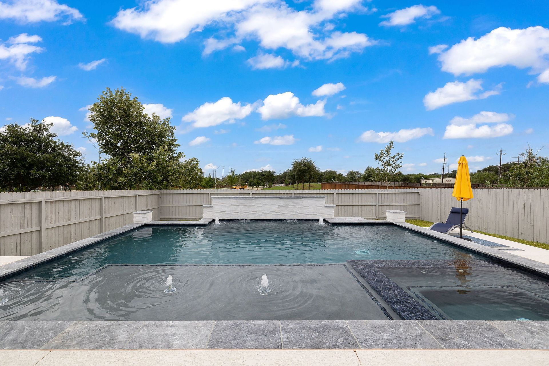 Backyard swimming pool with a concrete deck, white fence, and yellow umbrella under a blue sky