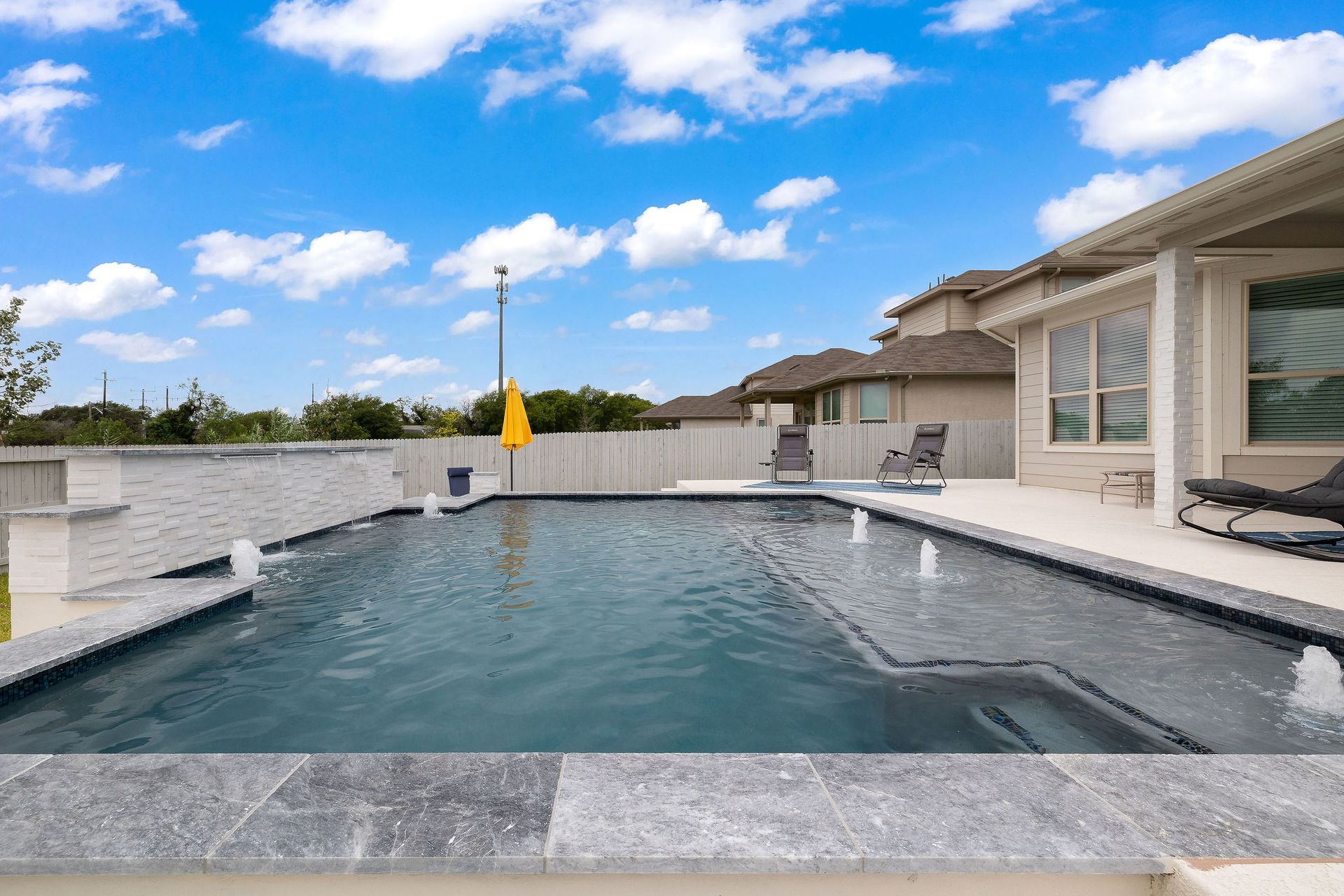 Modern backyard pool with spa, lounge chairs, and a patio under a bright blue sky
