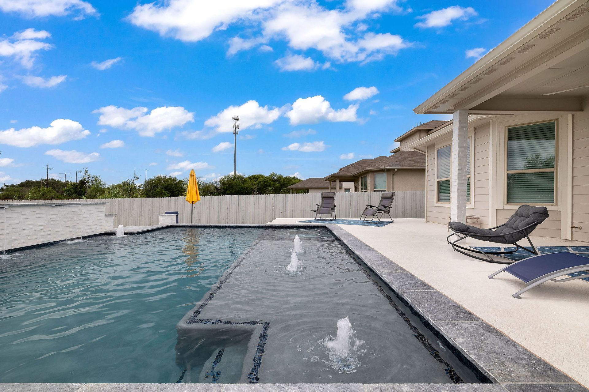Modern backyard pool with spa, lounge chairs, and a patio under a bright blue sky