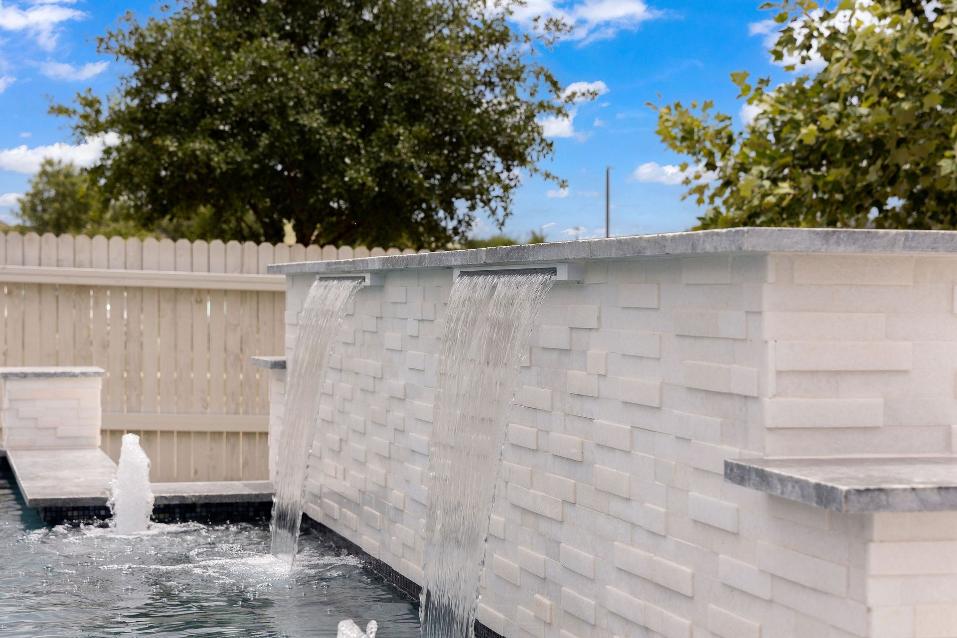 Modern white backyard fountain with cascading water and trees under a blue sky