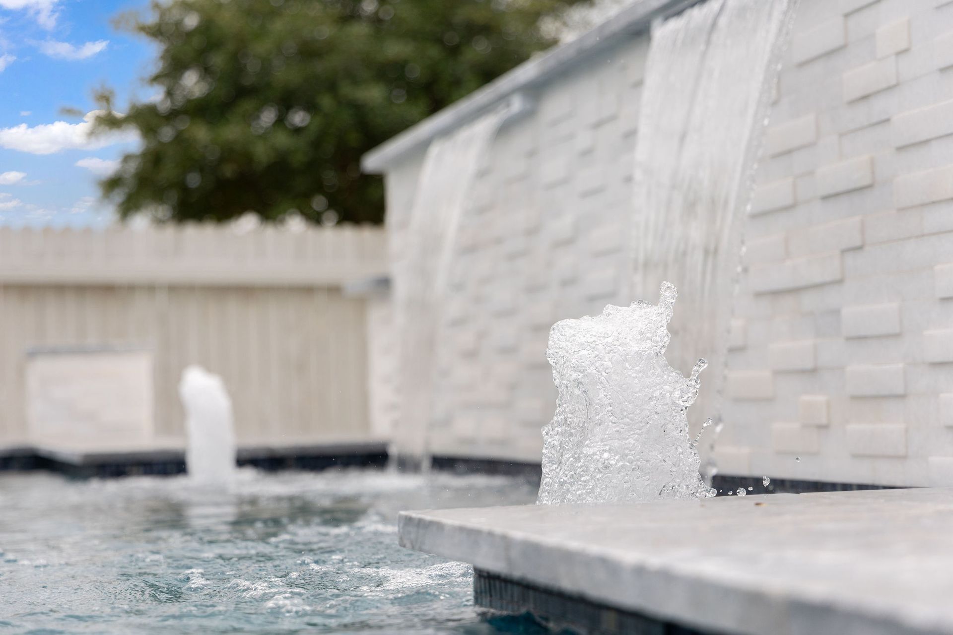 Small fountain jets splashing in a backyard pool beside a white house and fence