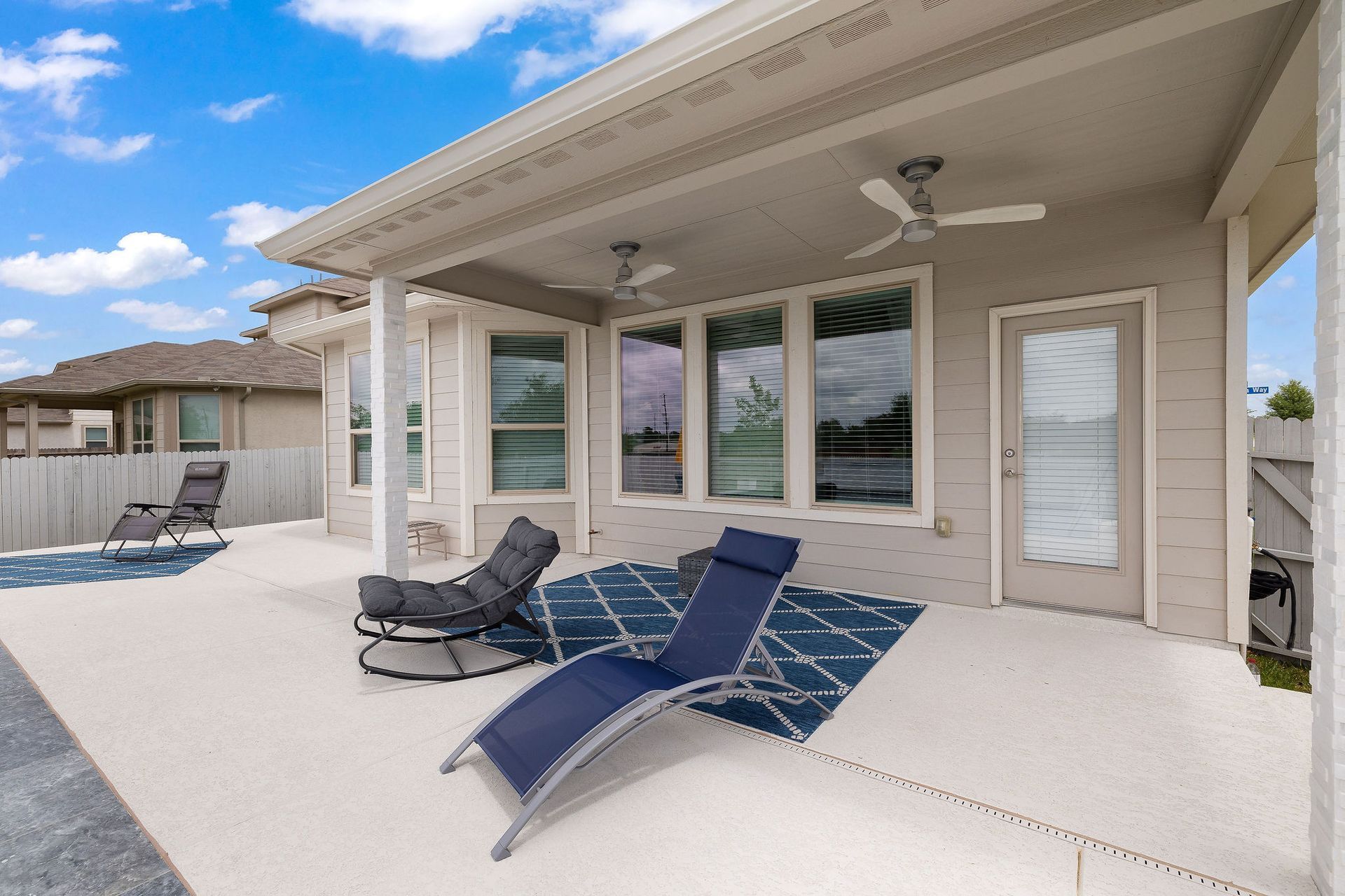 Covered patio with white lounge chairs, a pool, and sliding glass doors on a beige house