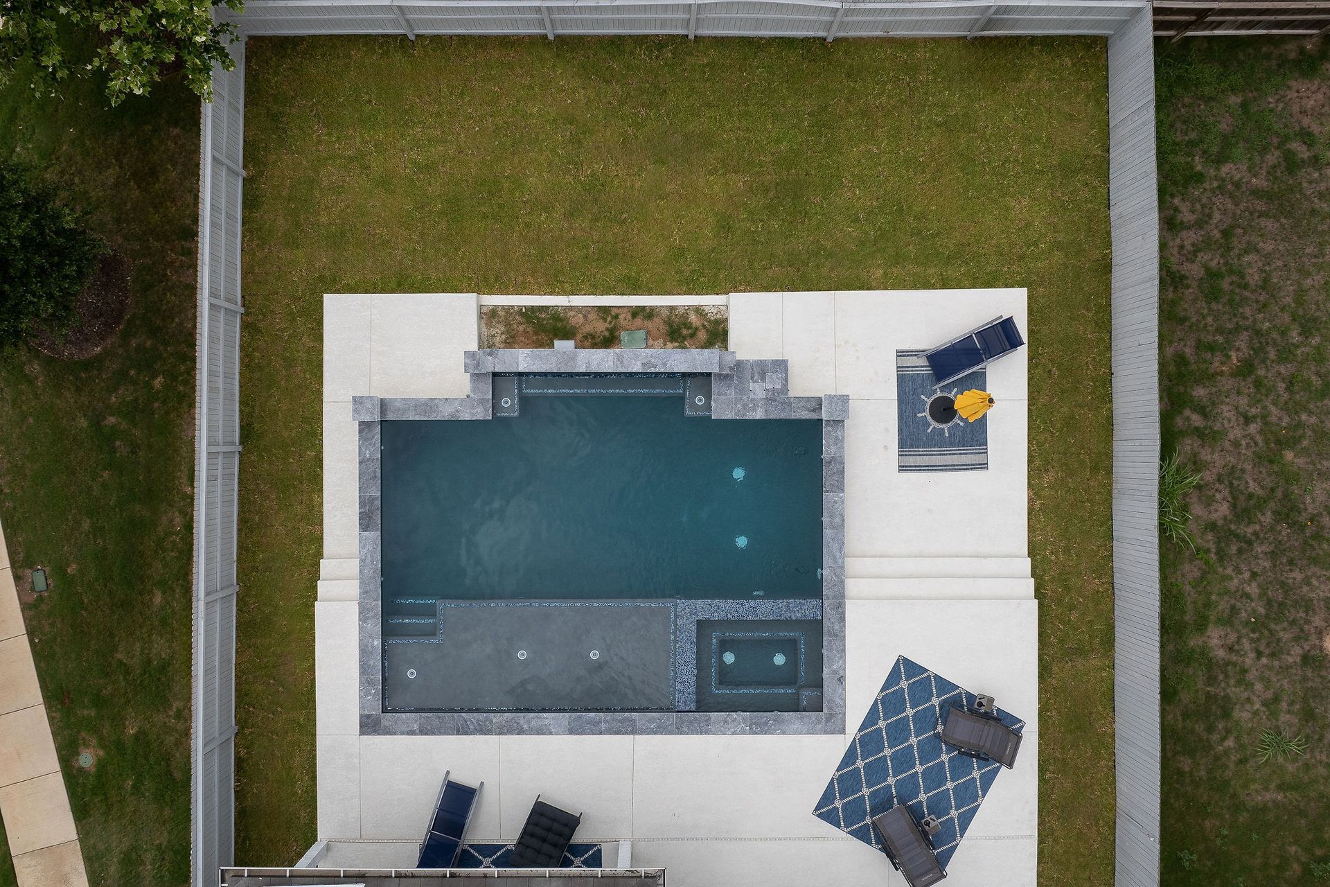 Top-down view of a backyard pool with lounge chairs and a patio table, surrounded by grass and concrete walls