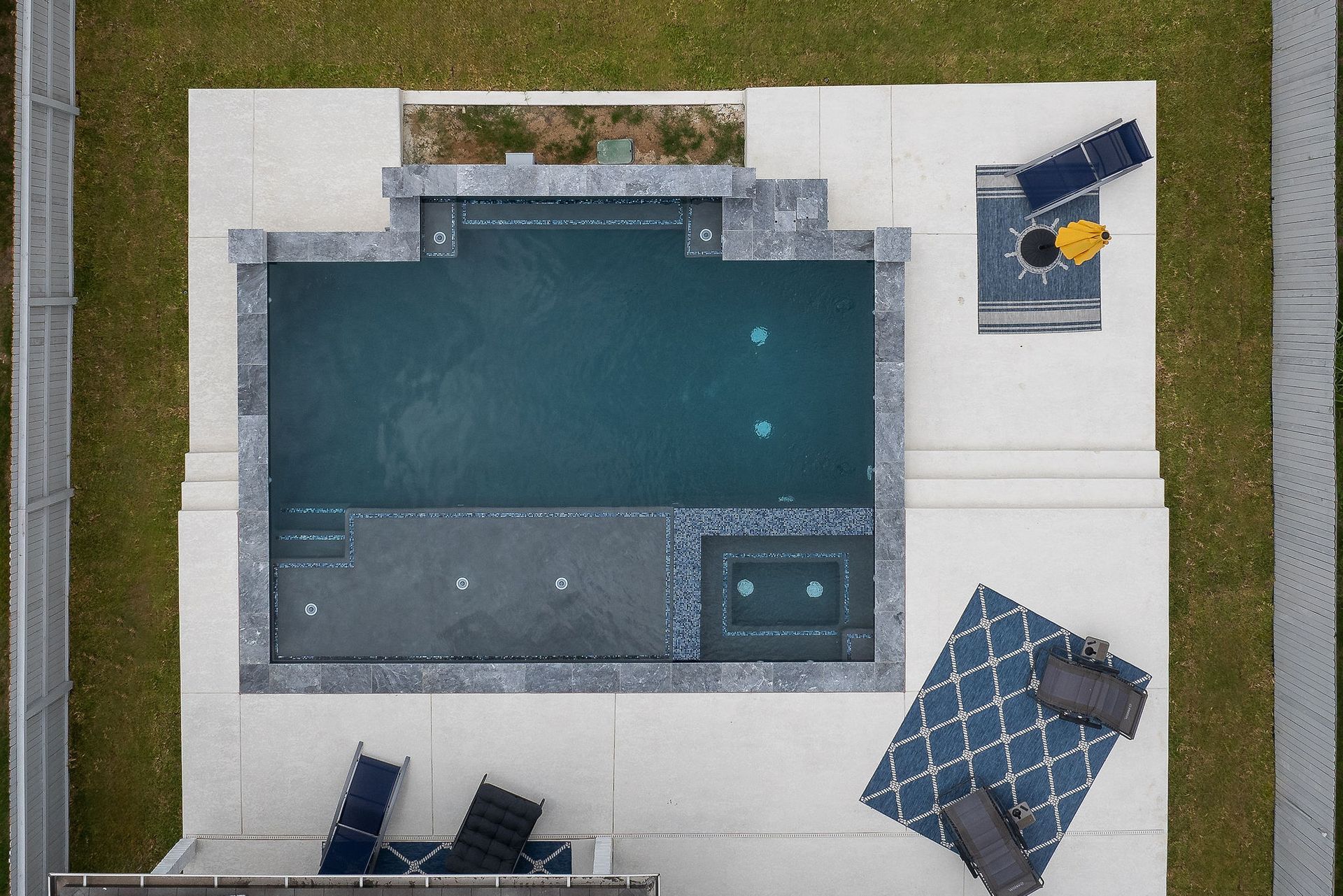 Top-down view of a modern backyard pool with white patio, dark loungers, and a blue patterned umbrella