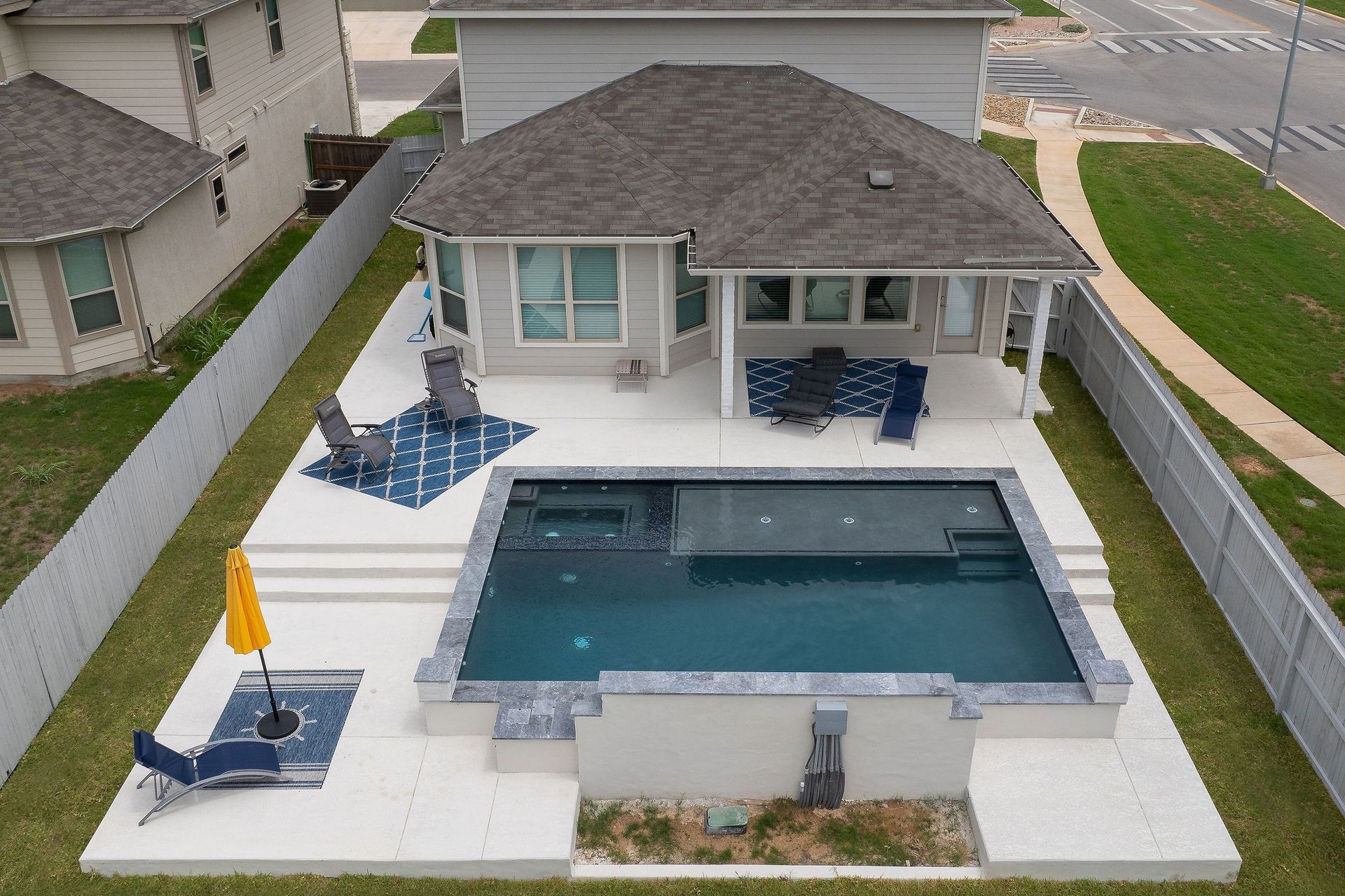 Backyard pool with patio seating beside a house, surrounded by a white fence and lawn