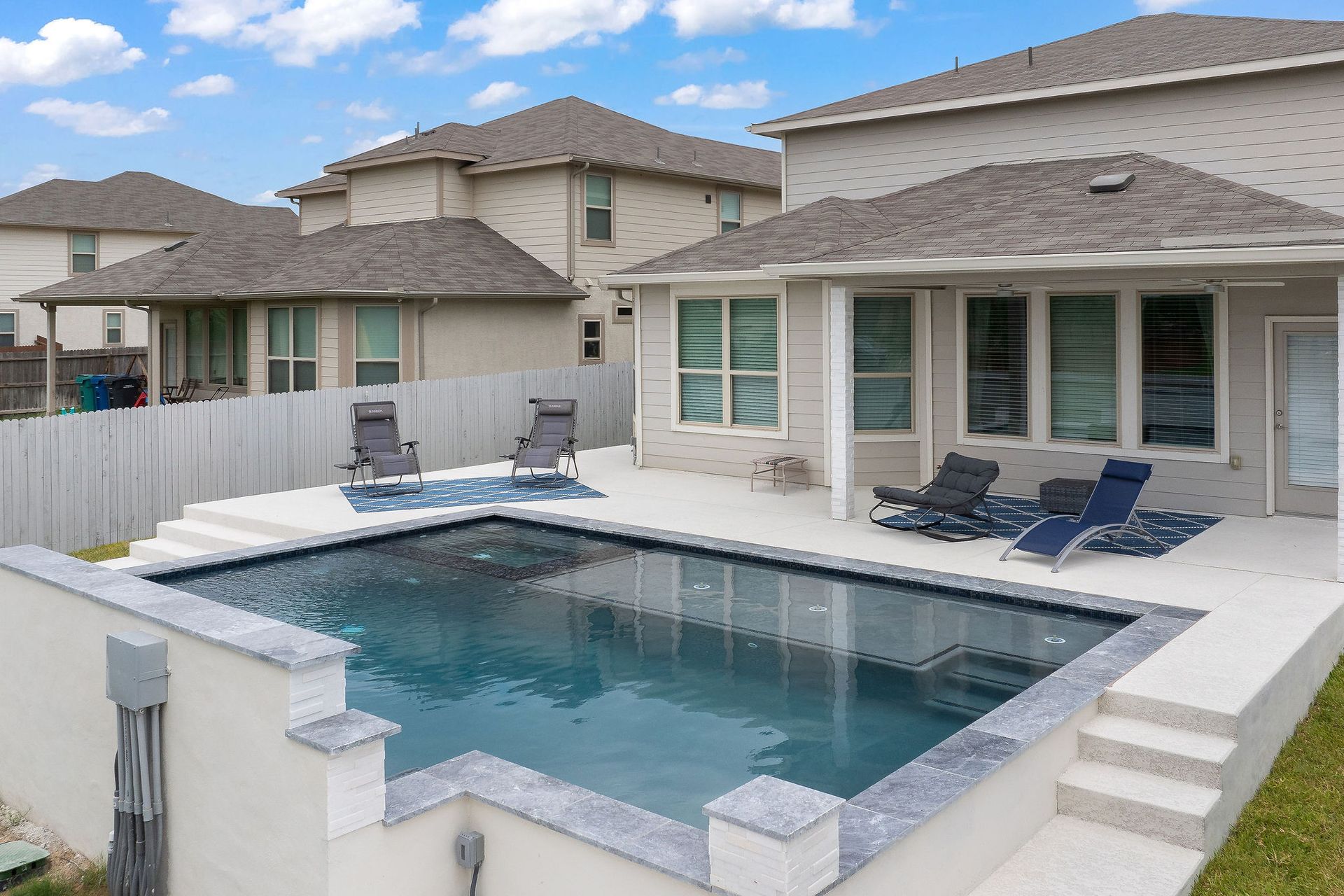 Backyard swimming pool with white patio, lounge chairs, and a house with large windows