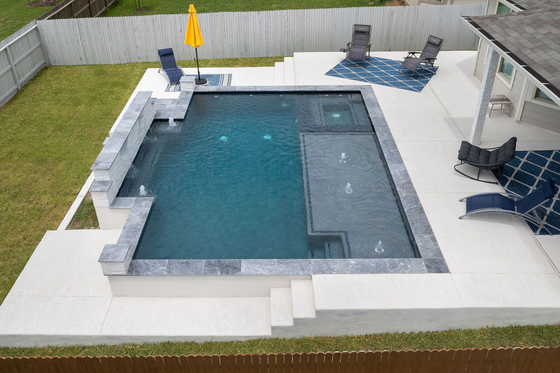 Backyard pool with gray stone deck, hot tub, patio chairs, and yellow umbrella