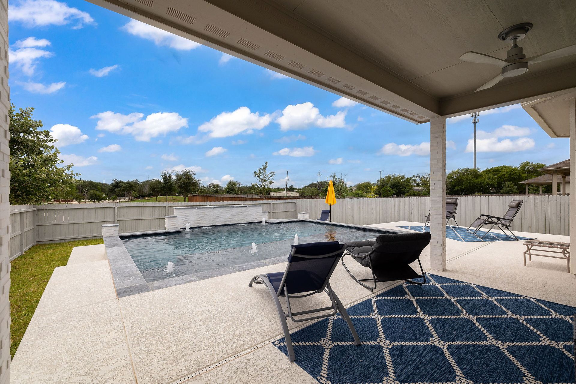 Covered patio with black lounge chairs and rug overlooking a backyard pool under a blue sky
