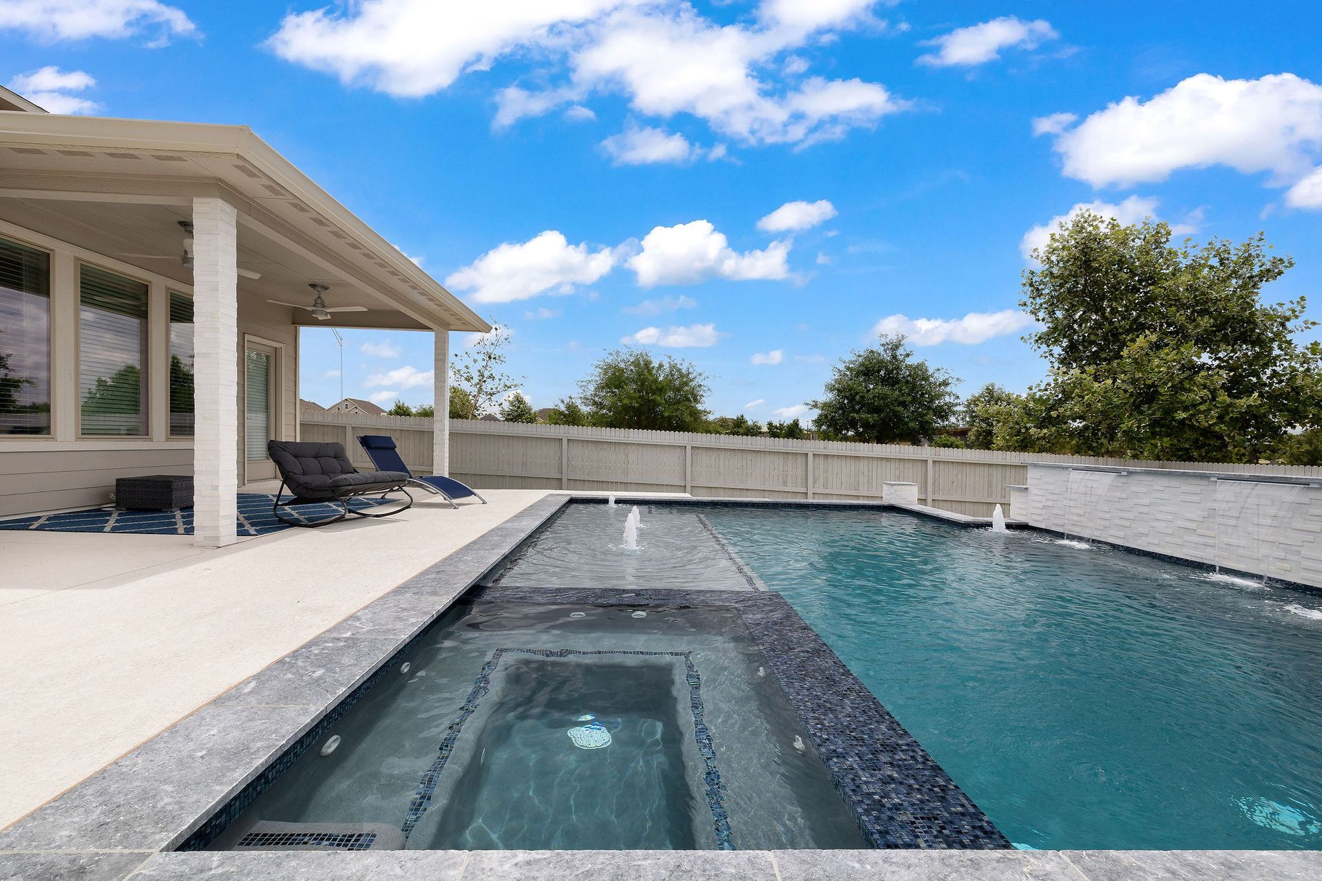 Modern backyard pool with a spa, patio chairs, and a covered patio under a blue sky