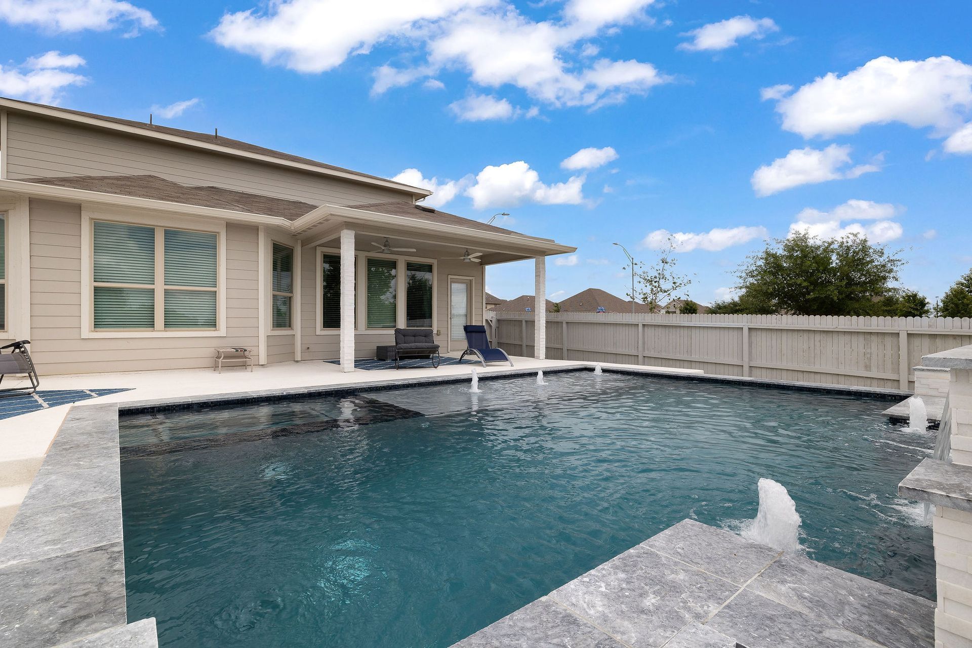 Backyard pool with stone deck beside a covered patio and white fence under a blue sky