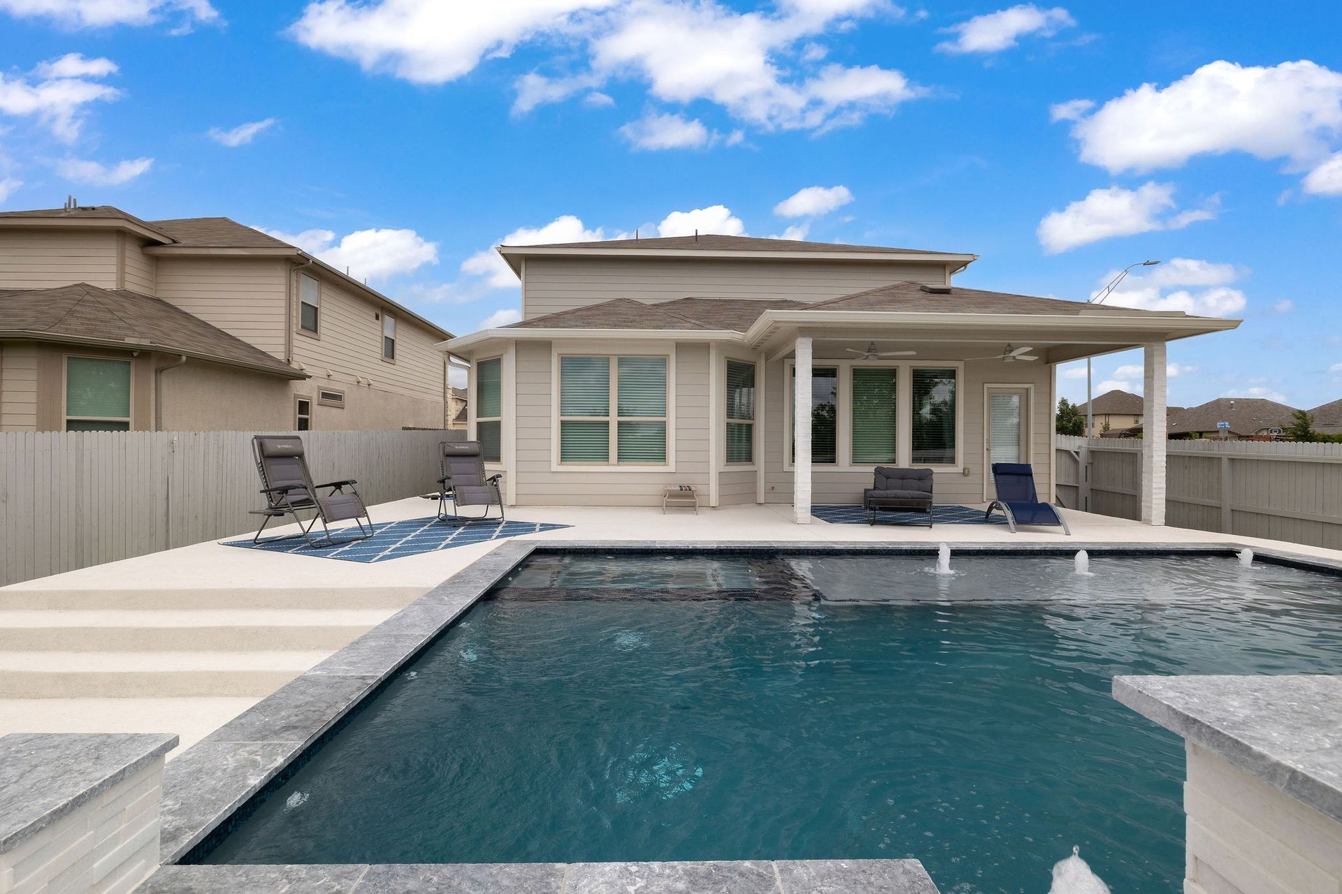 Modern backyard with a swimming pool, patio seating, and a two-story house under a blue sky