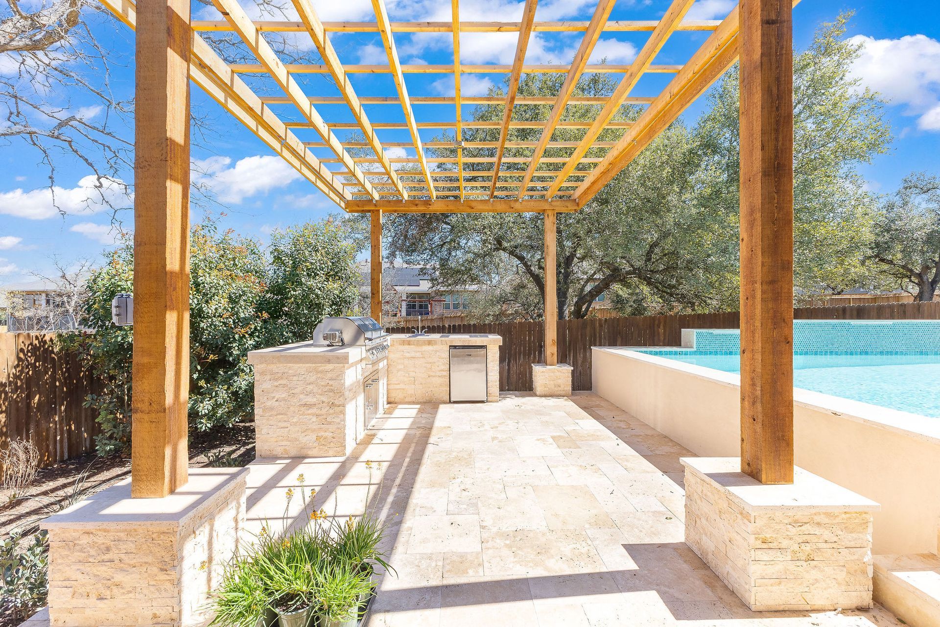 Wooden pergola over a stone patio with built-in grill, seating, and a pool in the background