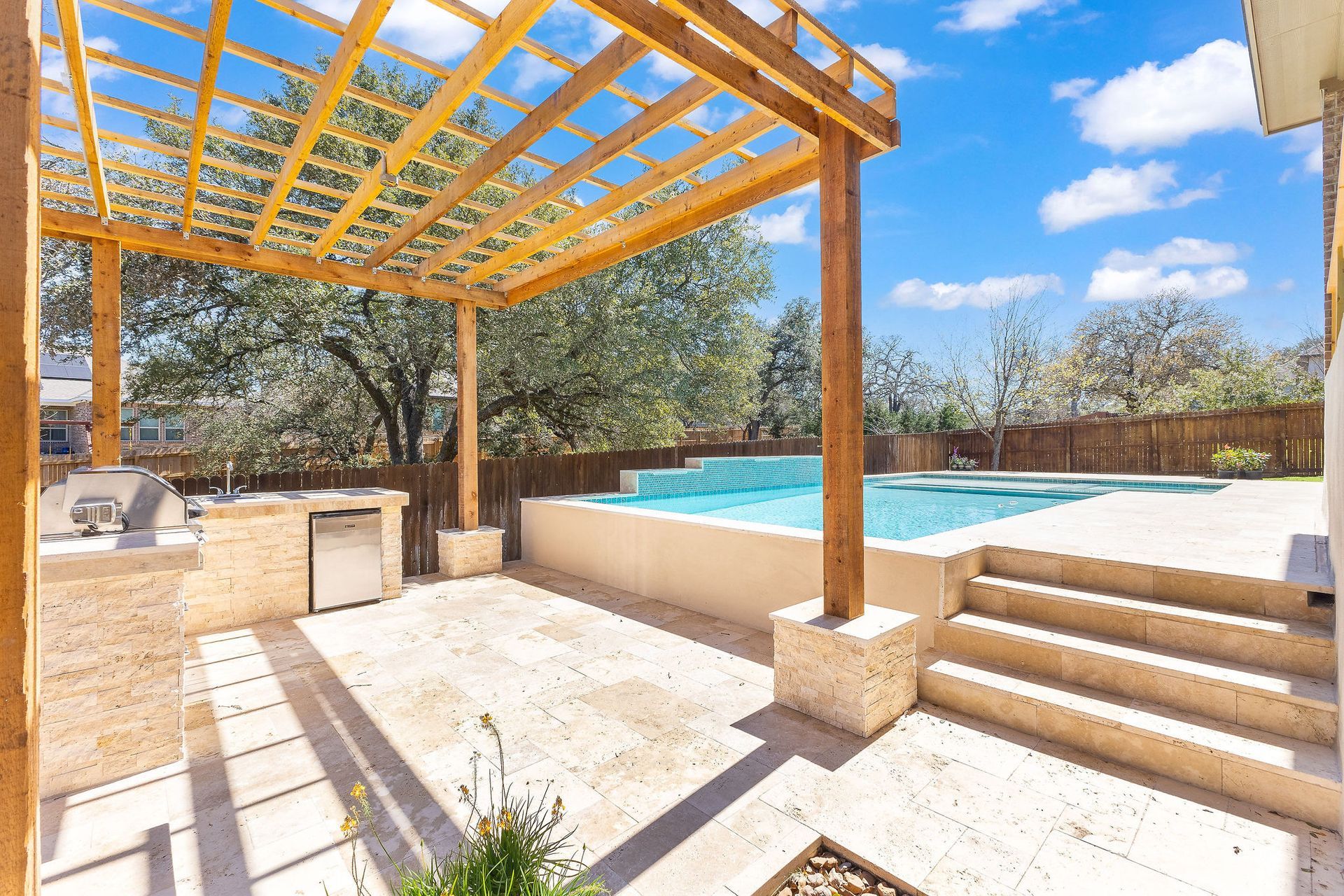Sunny backyard pool with wooden pergola, stone patio, and steps leading to the water