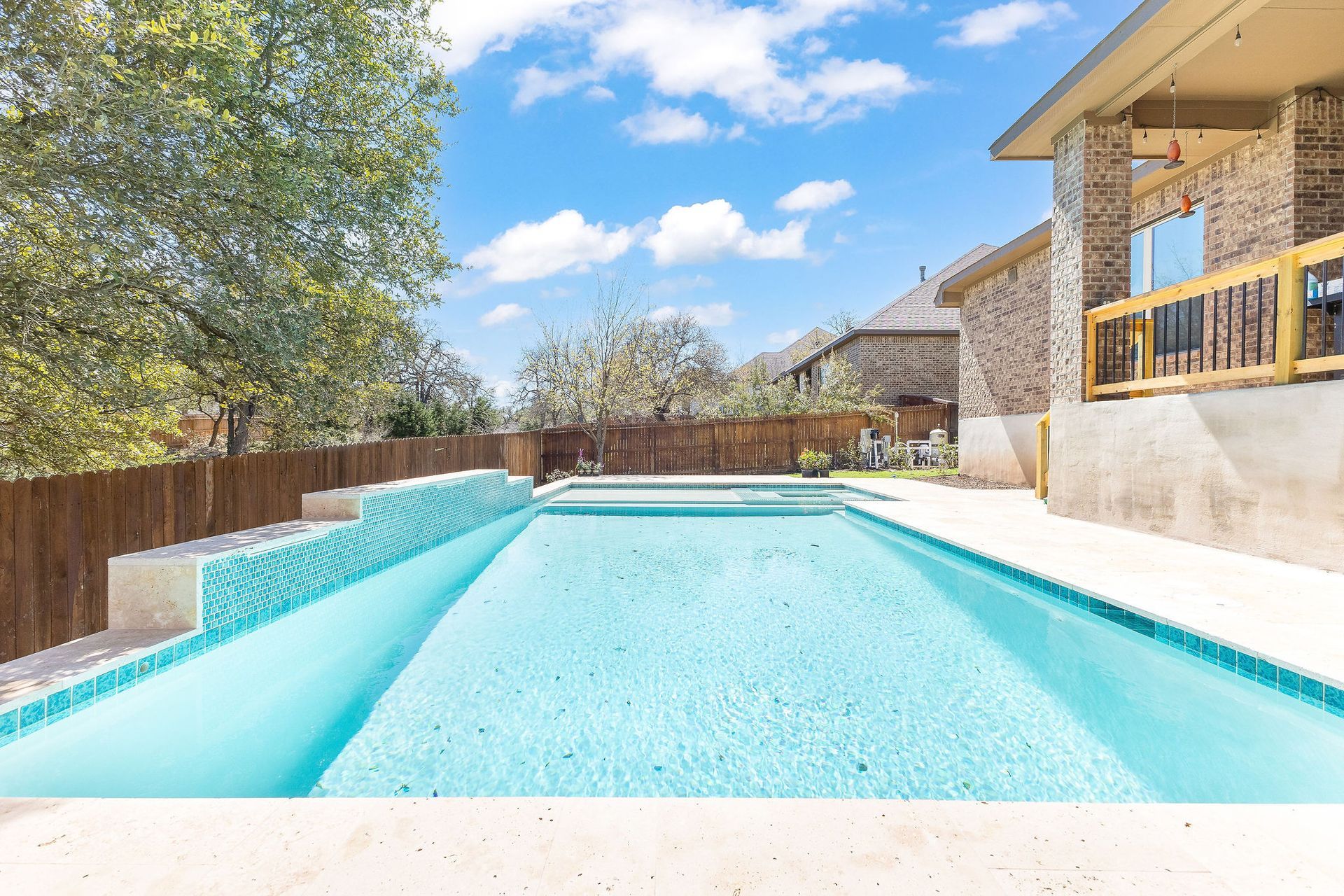 Backyard swimming pool with turquoise water beside a patio and trees under a bright blue sky