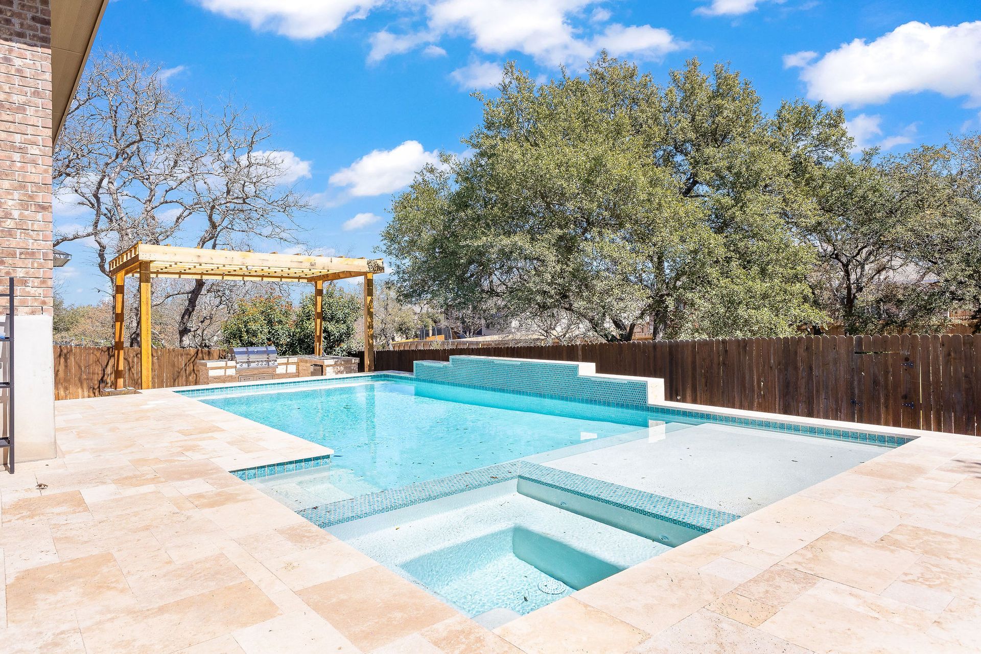 Backyard pool with spa and stone patio under a blue sky, bordered by trees and a wooden fence