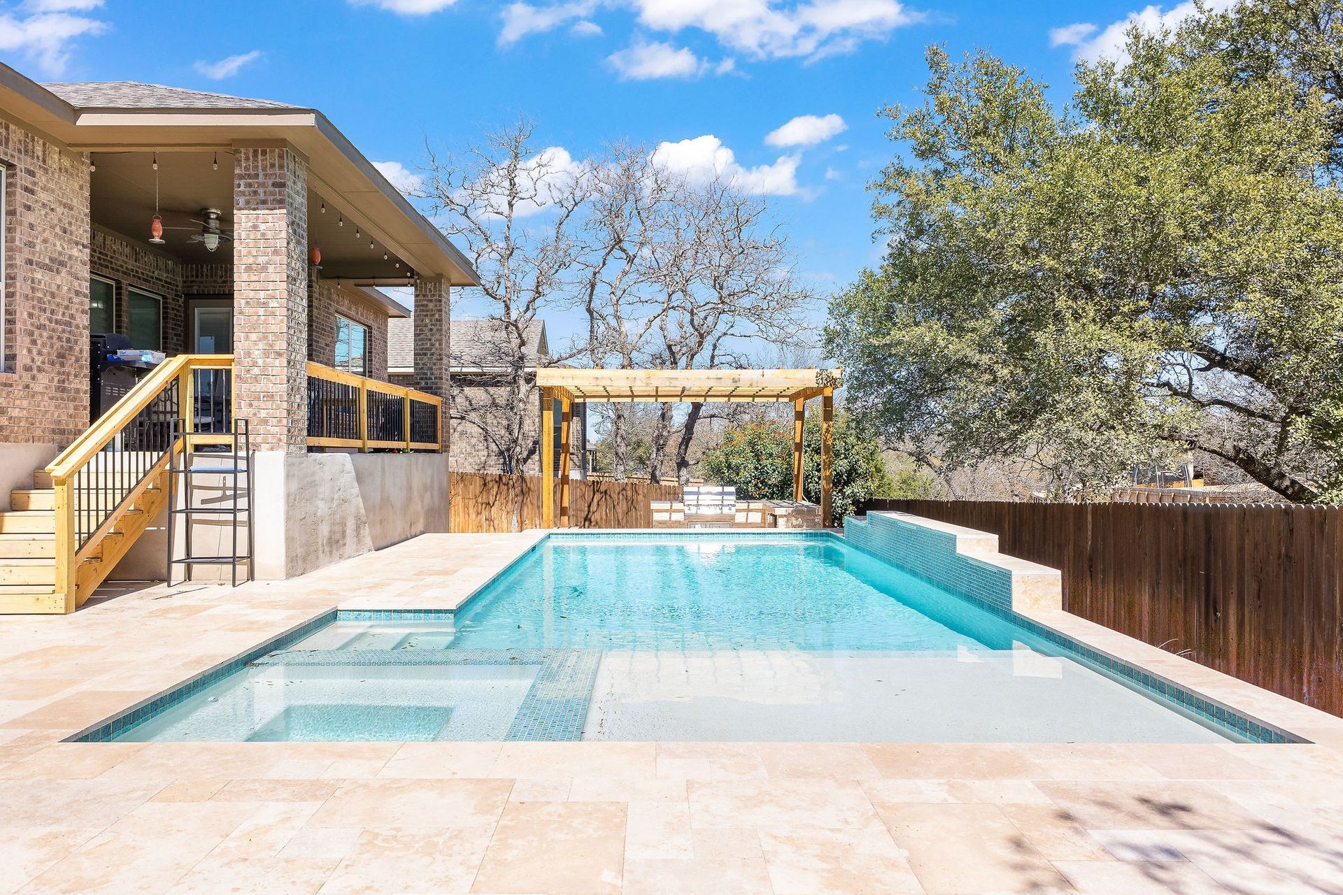 Backyard swimming pool beside a stone house, with a wooden pergola and trees under a blue sky.