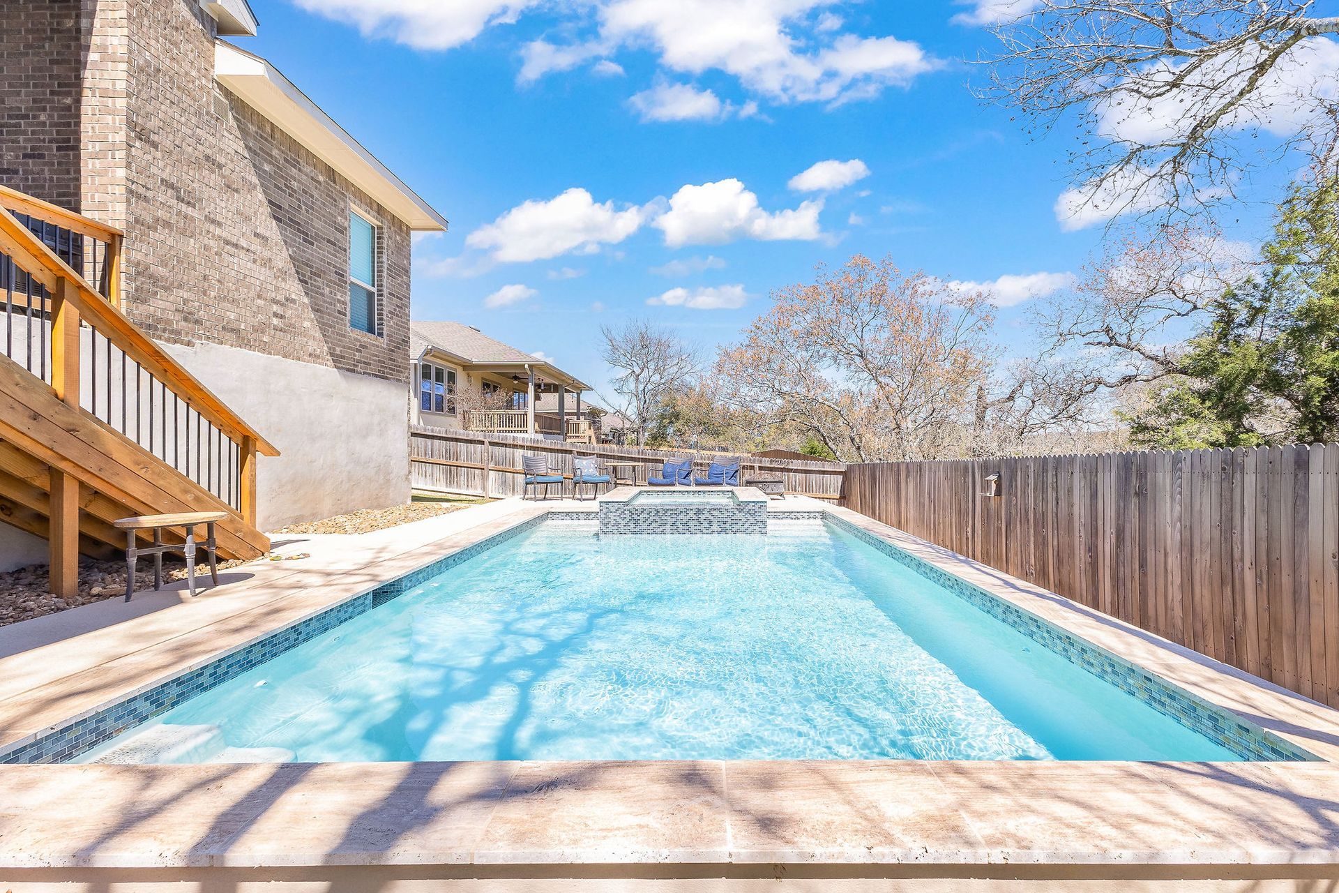 Backyard swimming pool beside a stone house and wooden fence under a bright blue sky
