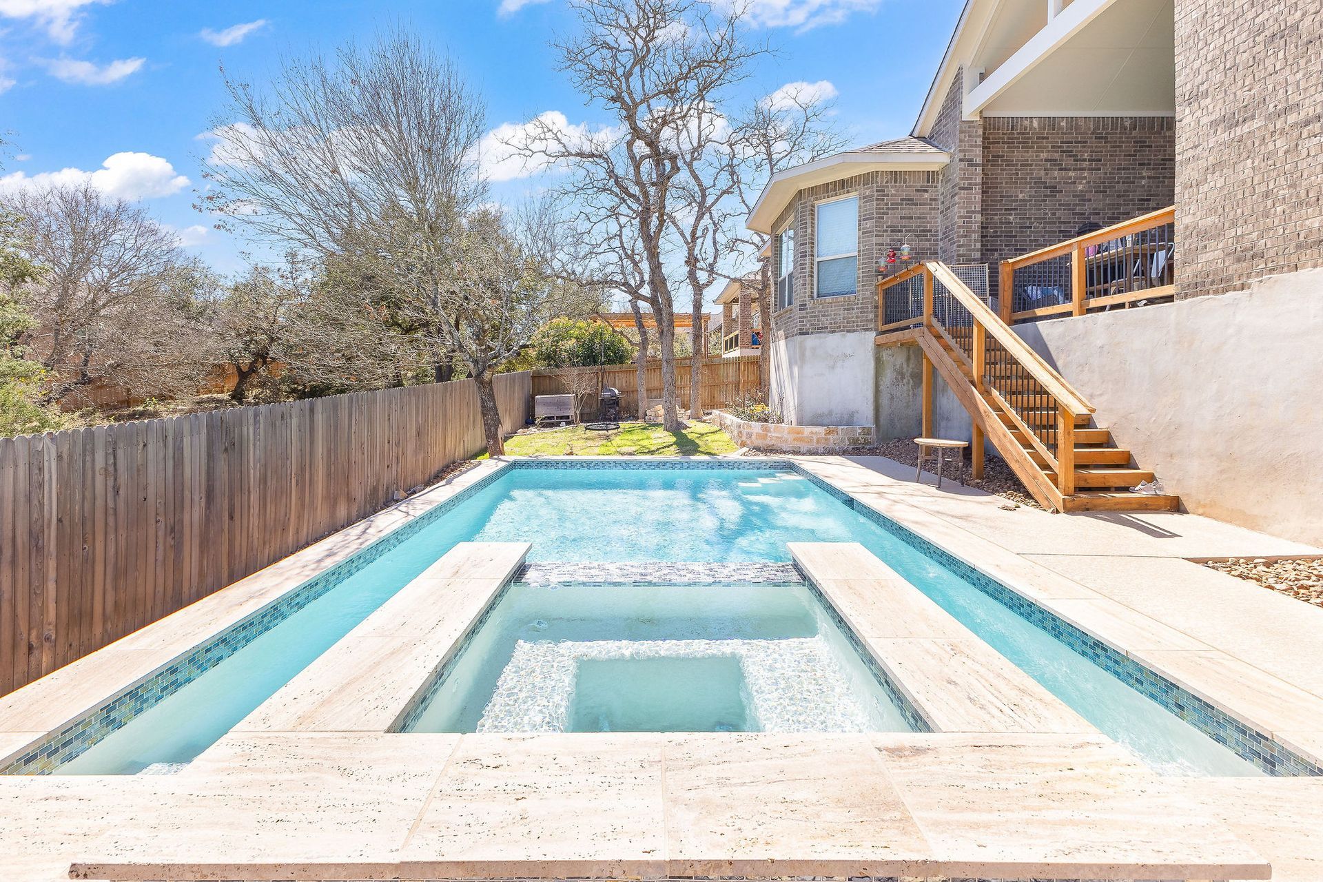 Backyard swimming pool with a raised spa, wooden fence, and house deck under a clear blue sky