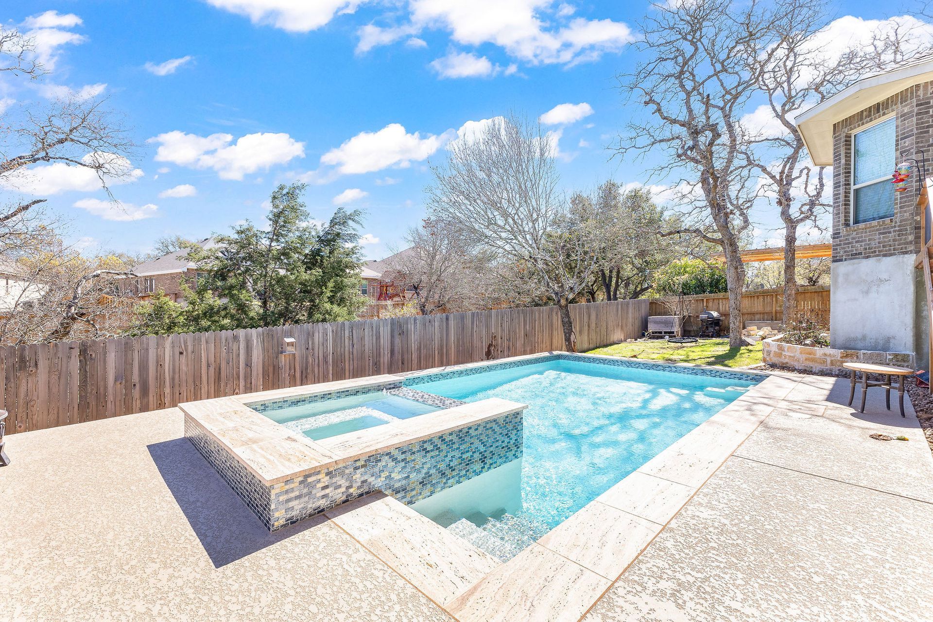 Backyard patio with a rectangular swimming pool, built-in spa, wooden fence, and trees under a blue sky