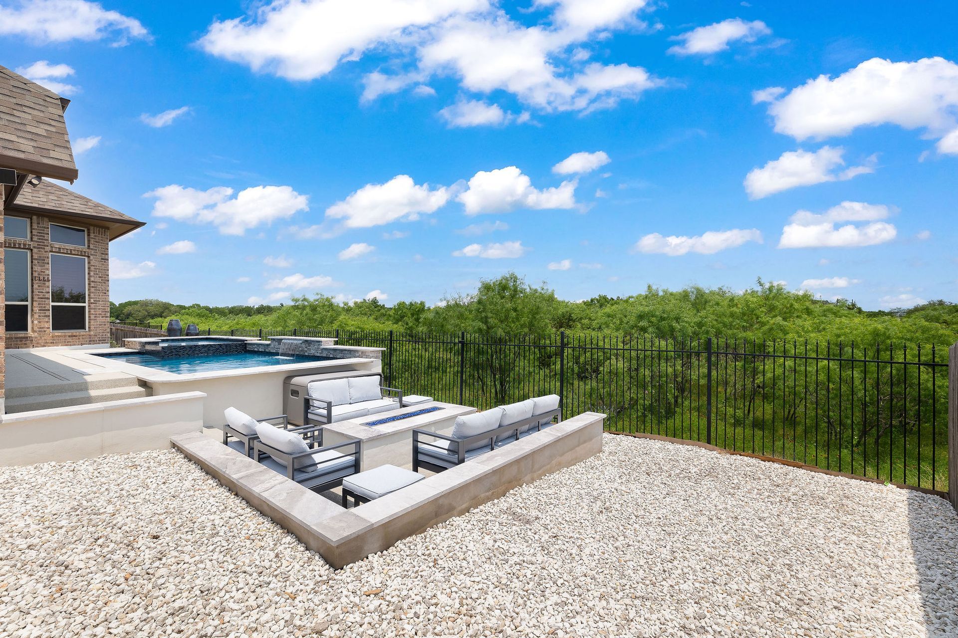 Modern backyard patio with outdoor sofa, fire pit, pool, and green trees under a blue sky