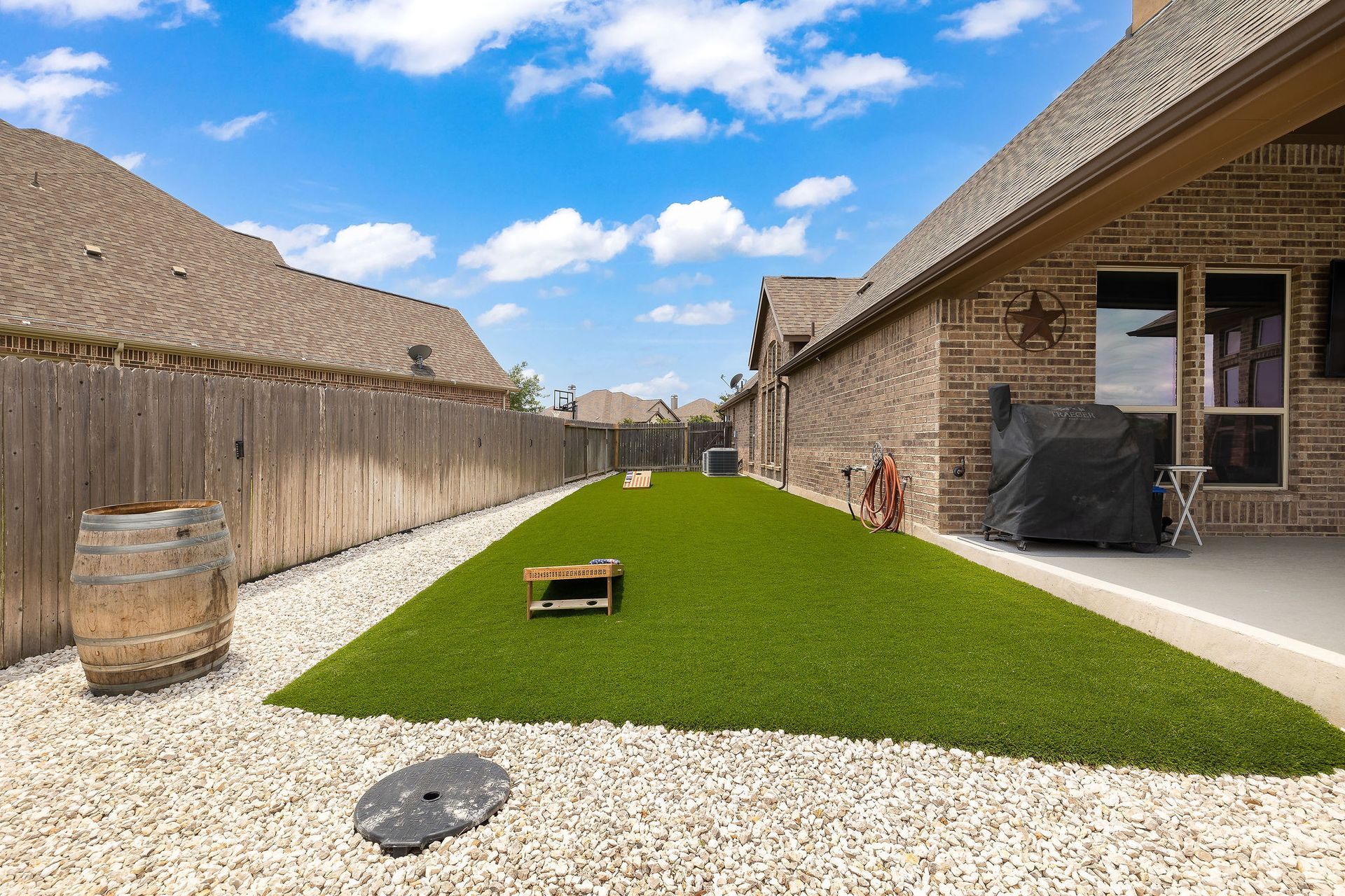 Backyard with artificial grass strip, gravel, wooden fence, and brick house under a blue sky