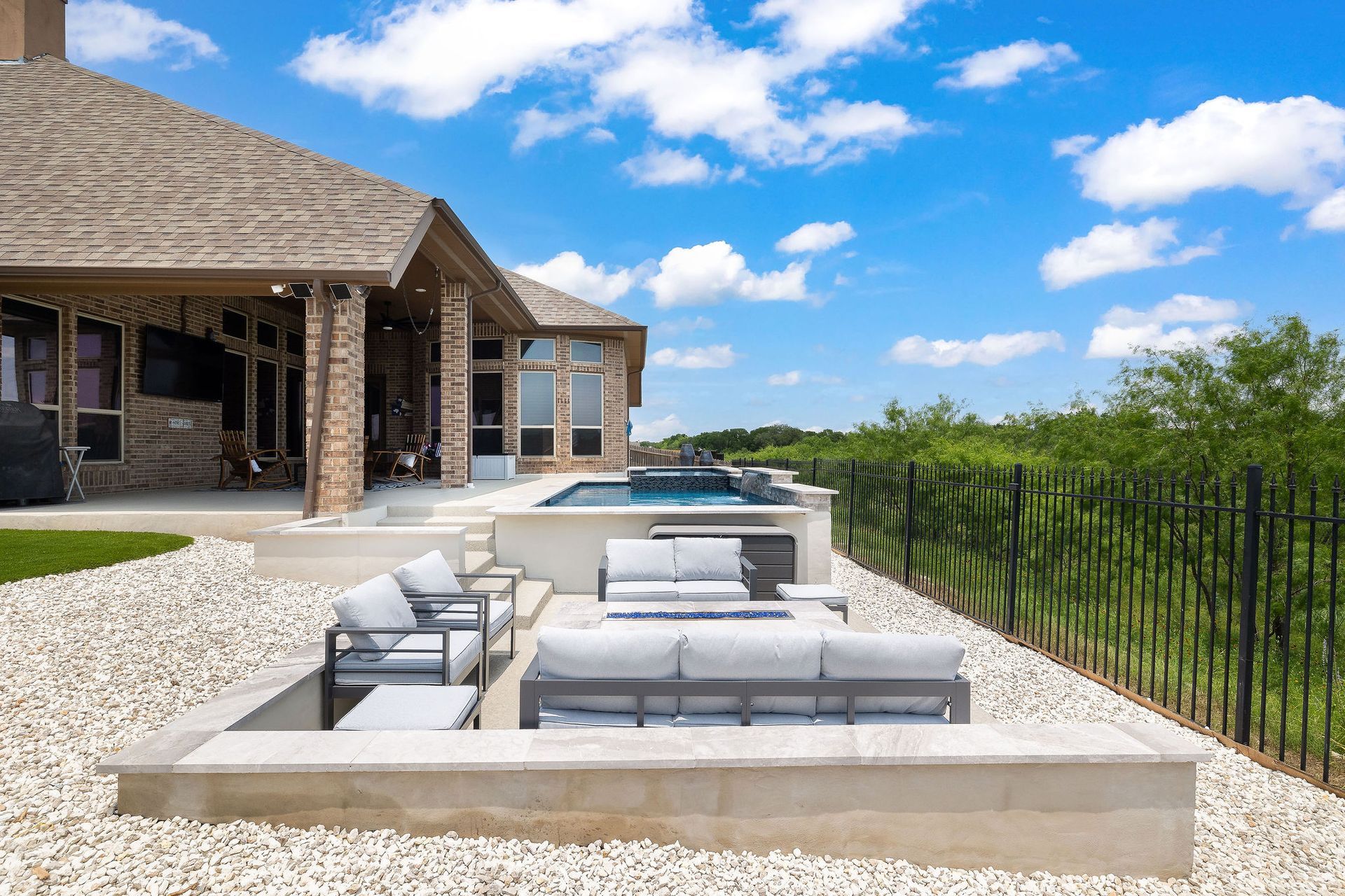 Outdoor patio with white lounge seating beside a stone house, overlooking a fenced green landscape under a blue sky