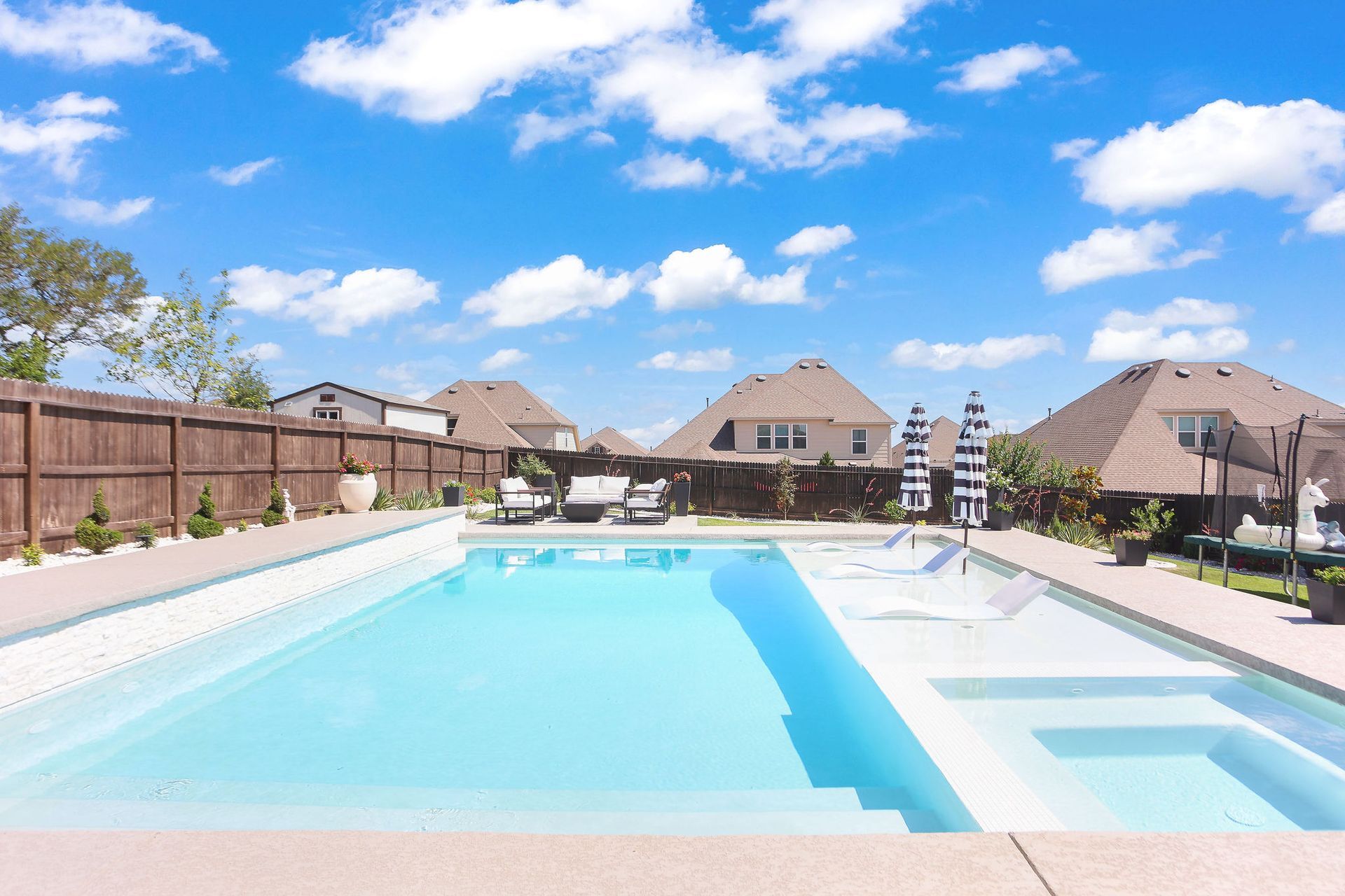 Sunny outdoor swimming pool with lounge chairs and umbrellas in a residential backyard.