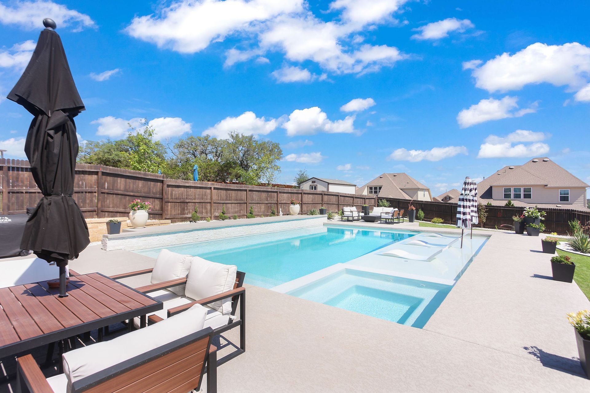 Sunny outdoor swimming pool with lounge chairs and umbrellas in a residential backyard.