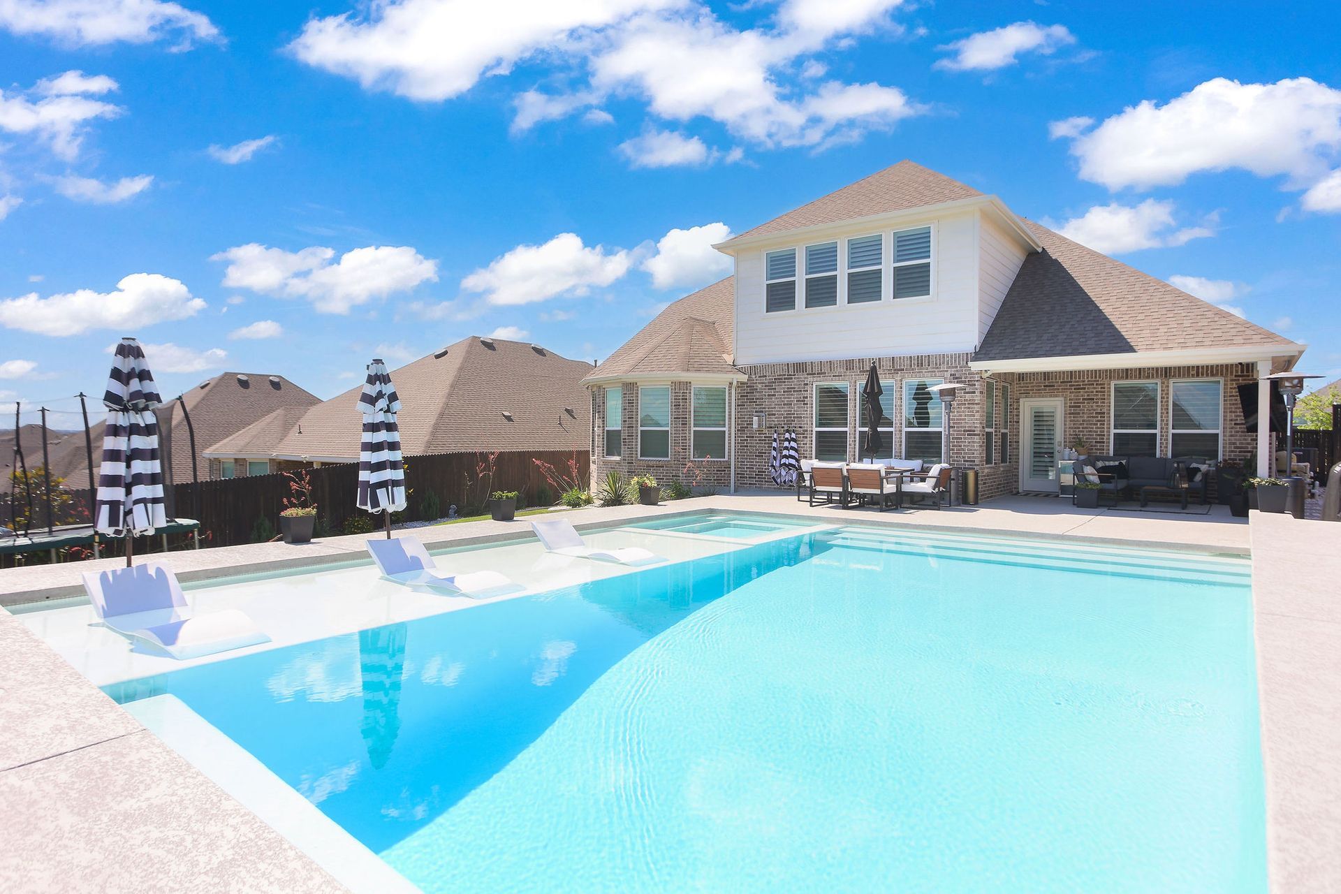 Sunny outdoor swimming pool with lounge chairs and umbrellas in a residential backyard.