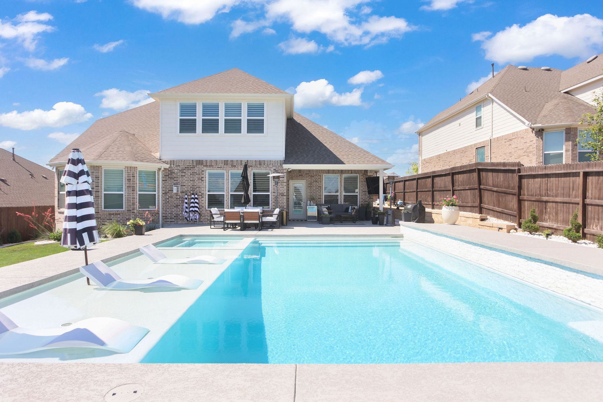 Sunny outdoor swimming pool with lounge chairs and umbrellas in a residential backyard.