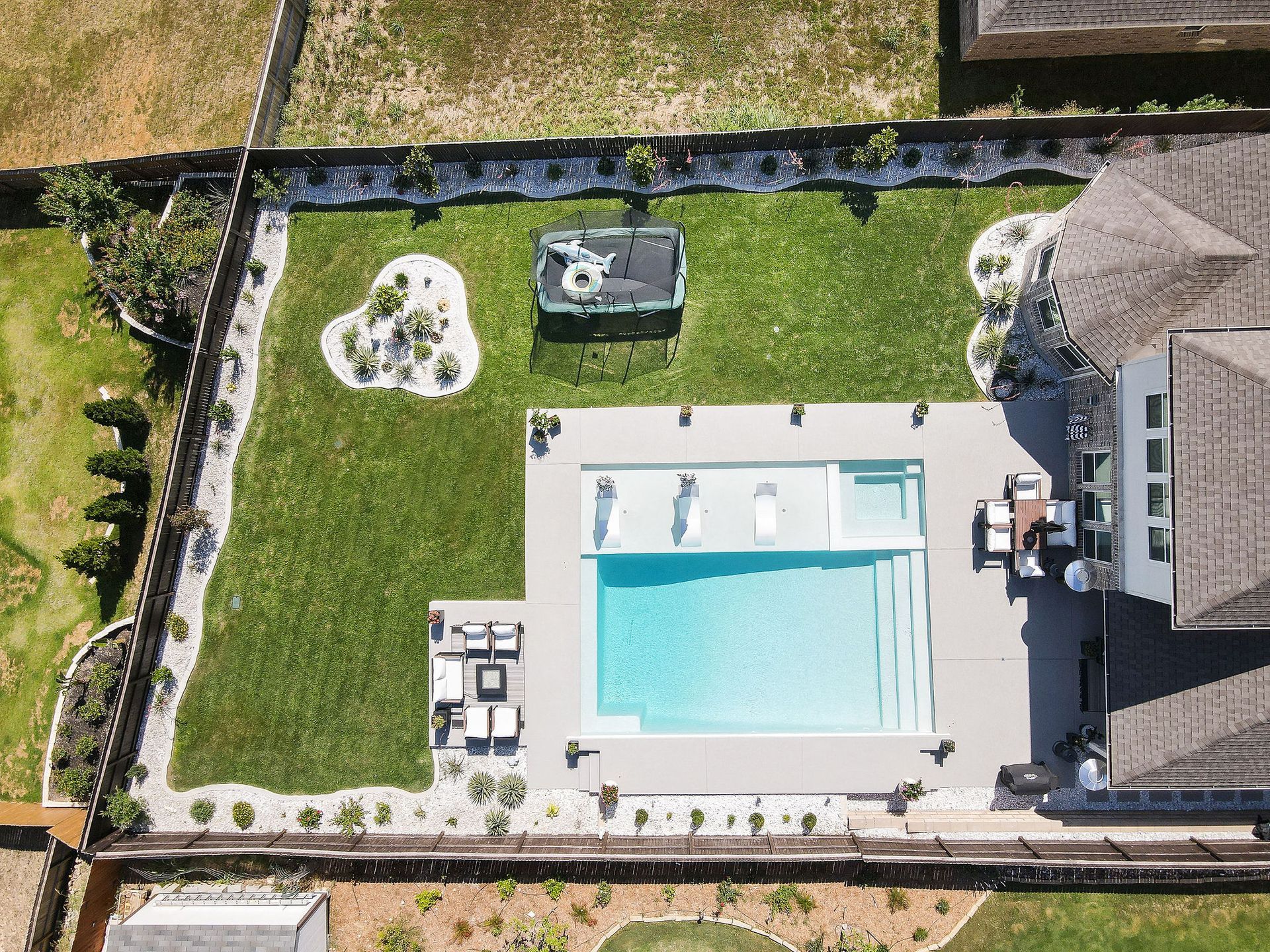 Aerial view of a backyard pool with lounge chairs, patio seating, and a grassy yard enclosed by a black fence