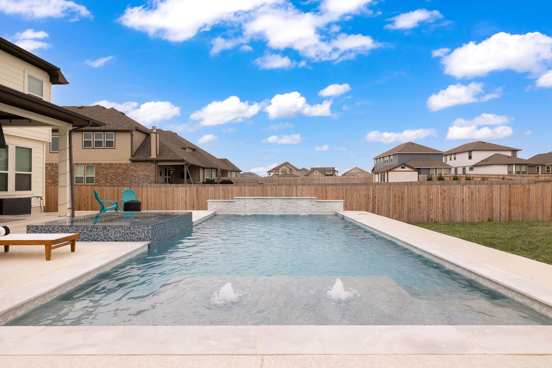 Backyard swimming pool with stone deck, wooden fence, and suburban houses under a blue sky