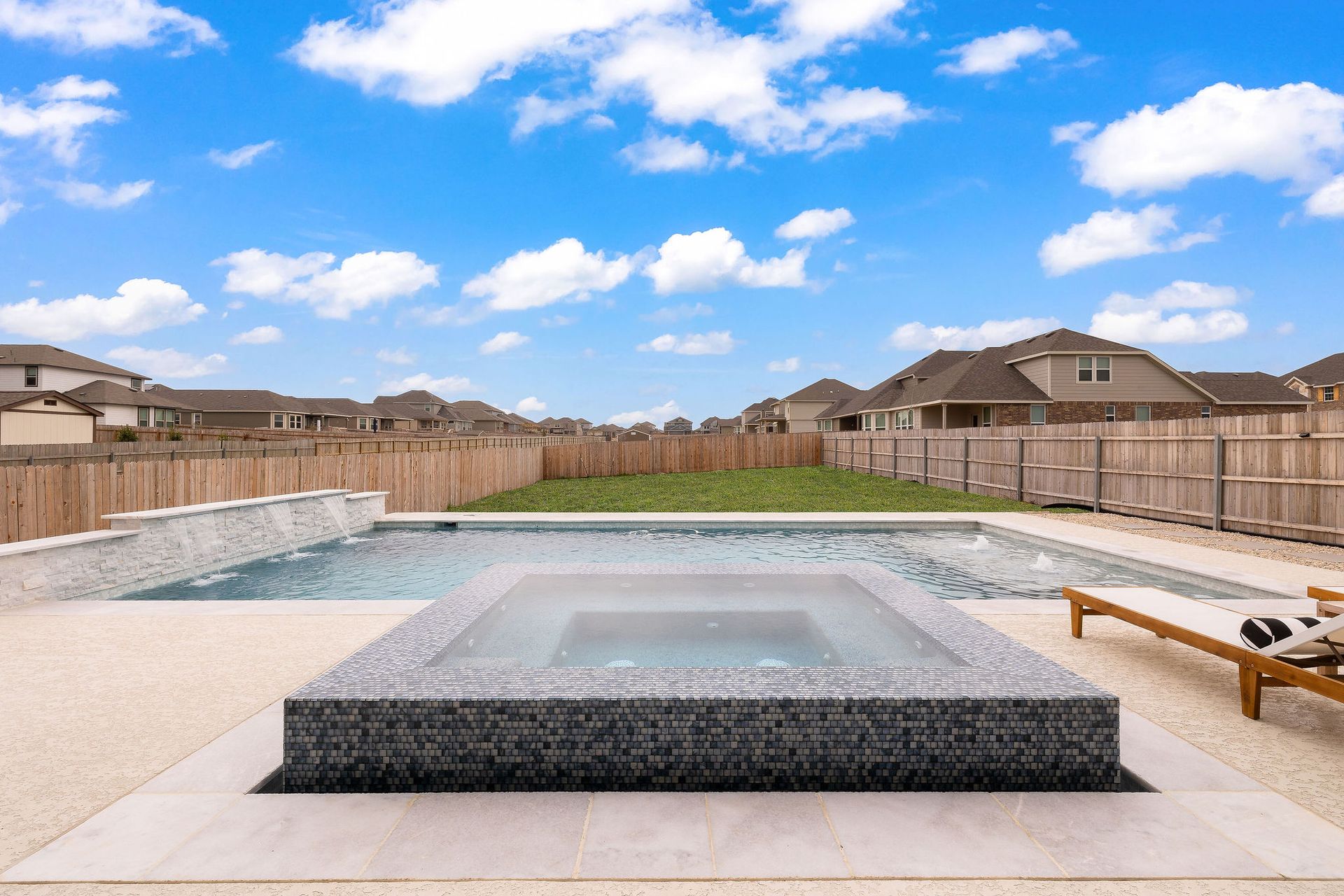 Backyard with a rectangular pool, raised spa, lounge chairs, and fenced suburban homes under a blue sky