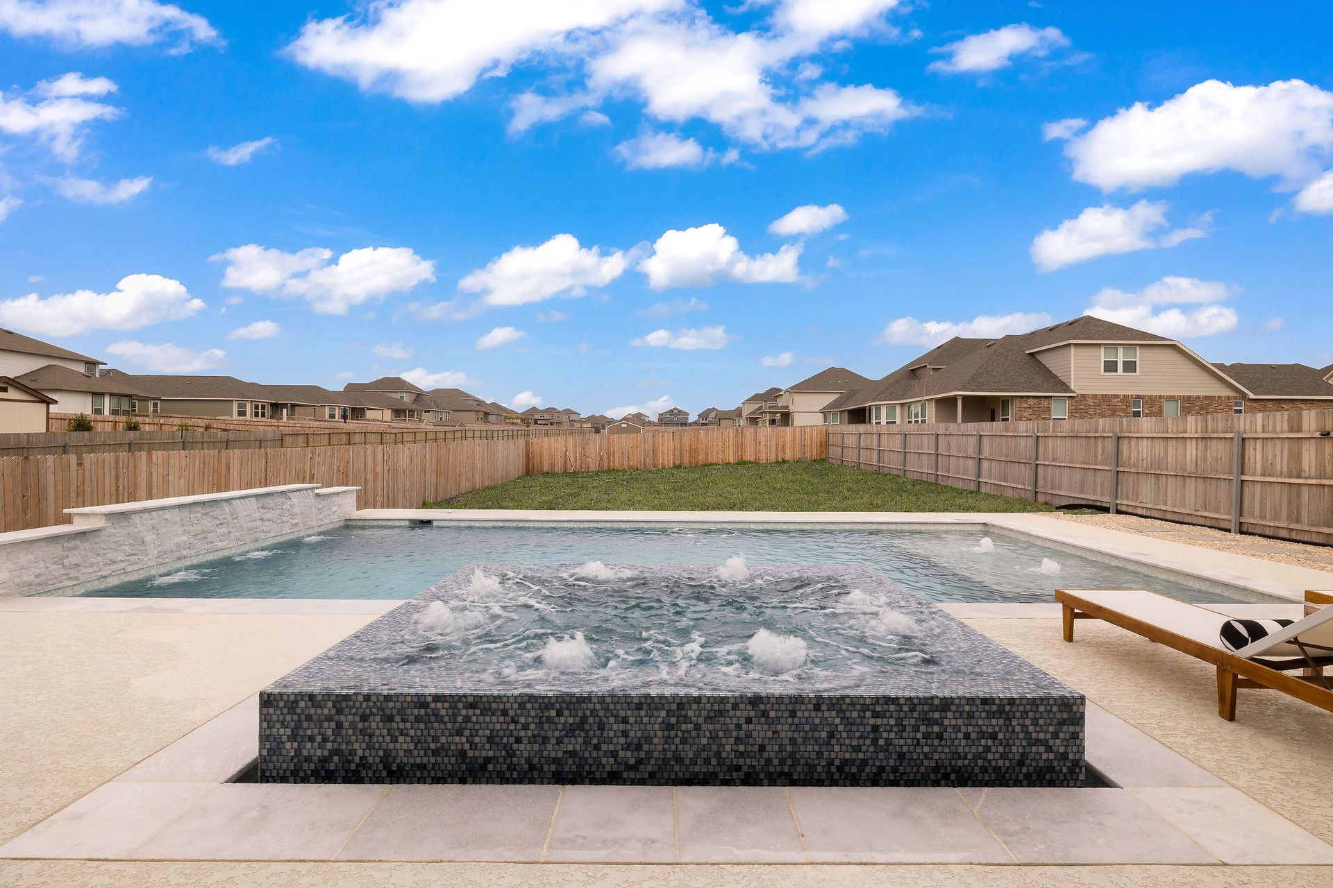 Backyard with a rectangular pool, raised spa, lounge chairs, and fenced suburban homes under a blue sky