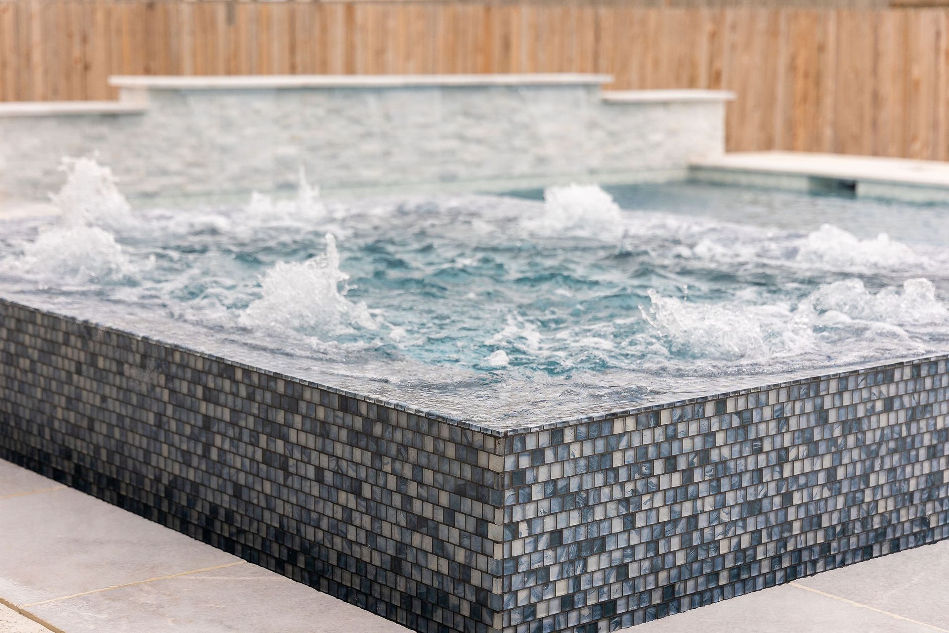 Modern backyard hot tub with bubbling water, stone tile exterior, and a wooden privacy fence behind it
