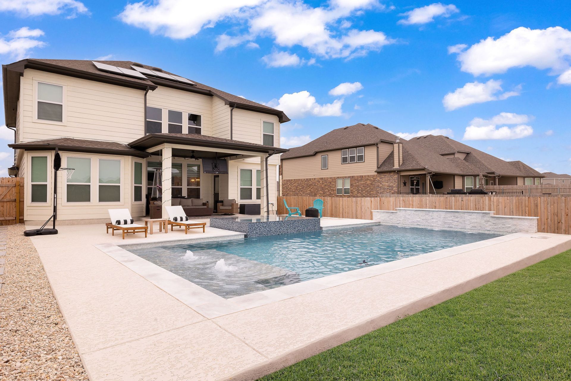 Backyard patio with a large swimming pool beside a two-story house under a blue sky