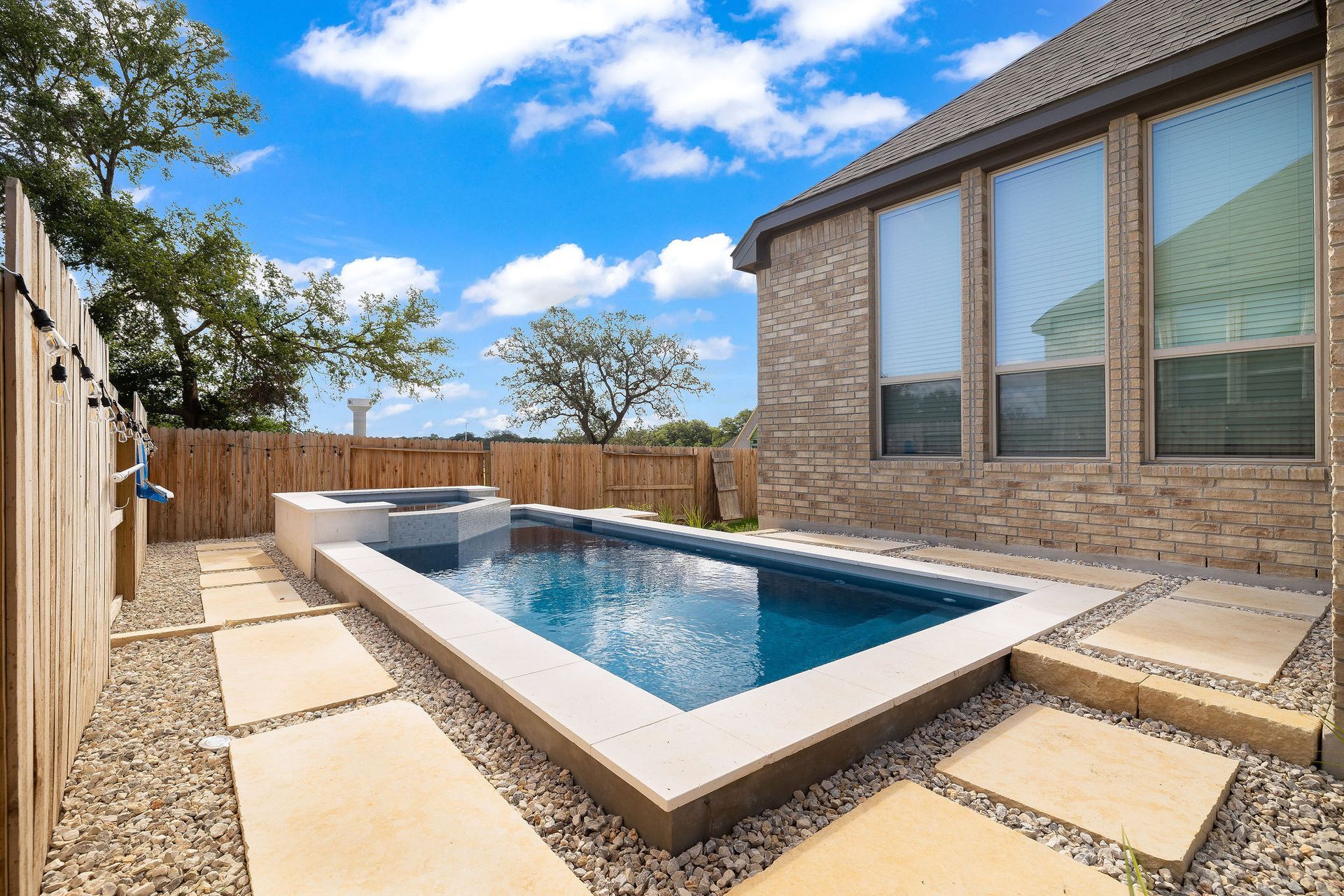 Backyard pool with stone deck beside a brick house under a blue sky