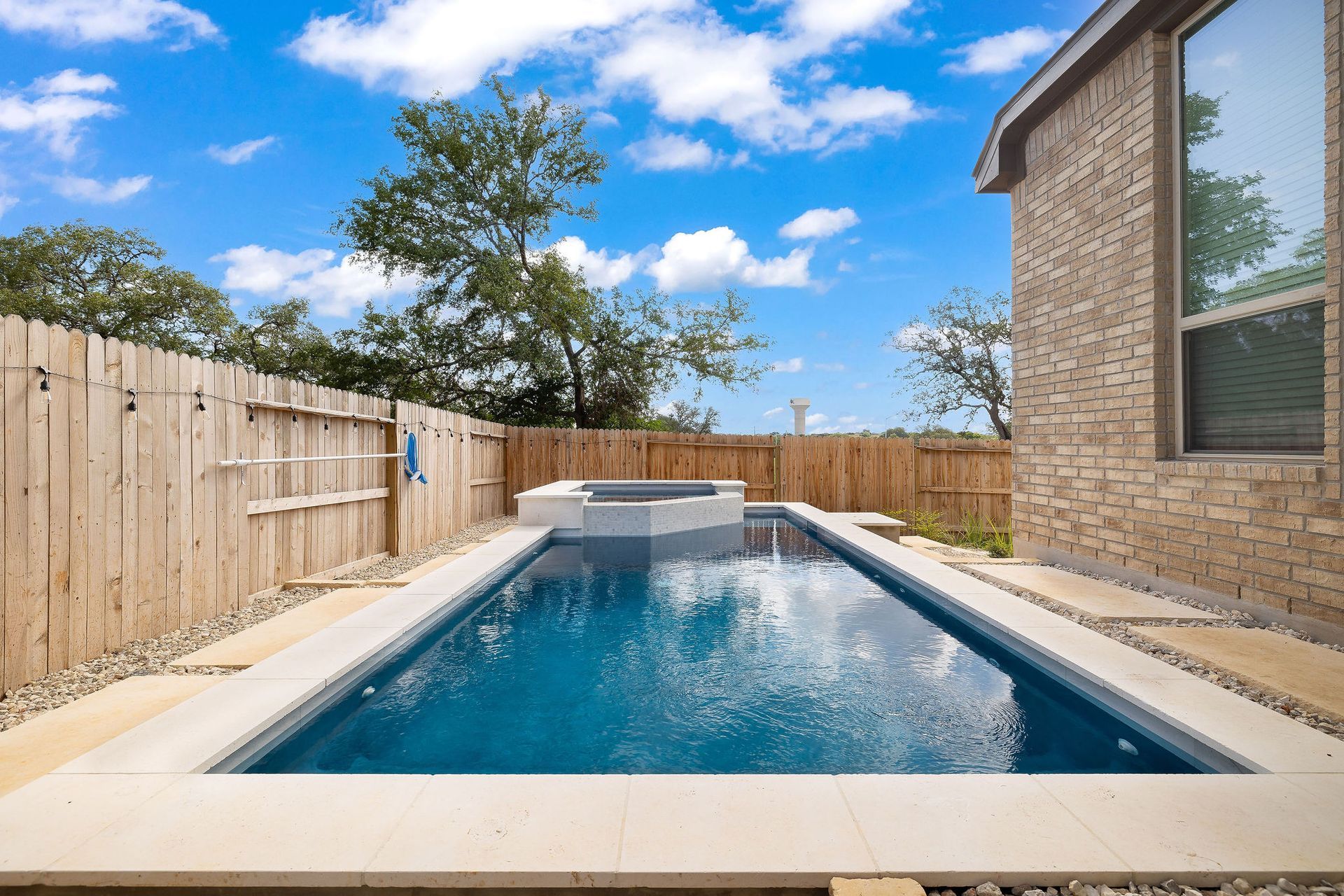 Backyard lap pool with blue water beside a brick house and wooden privacy fence under a sunny sky