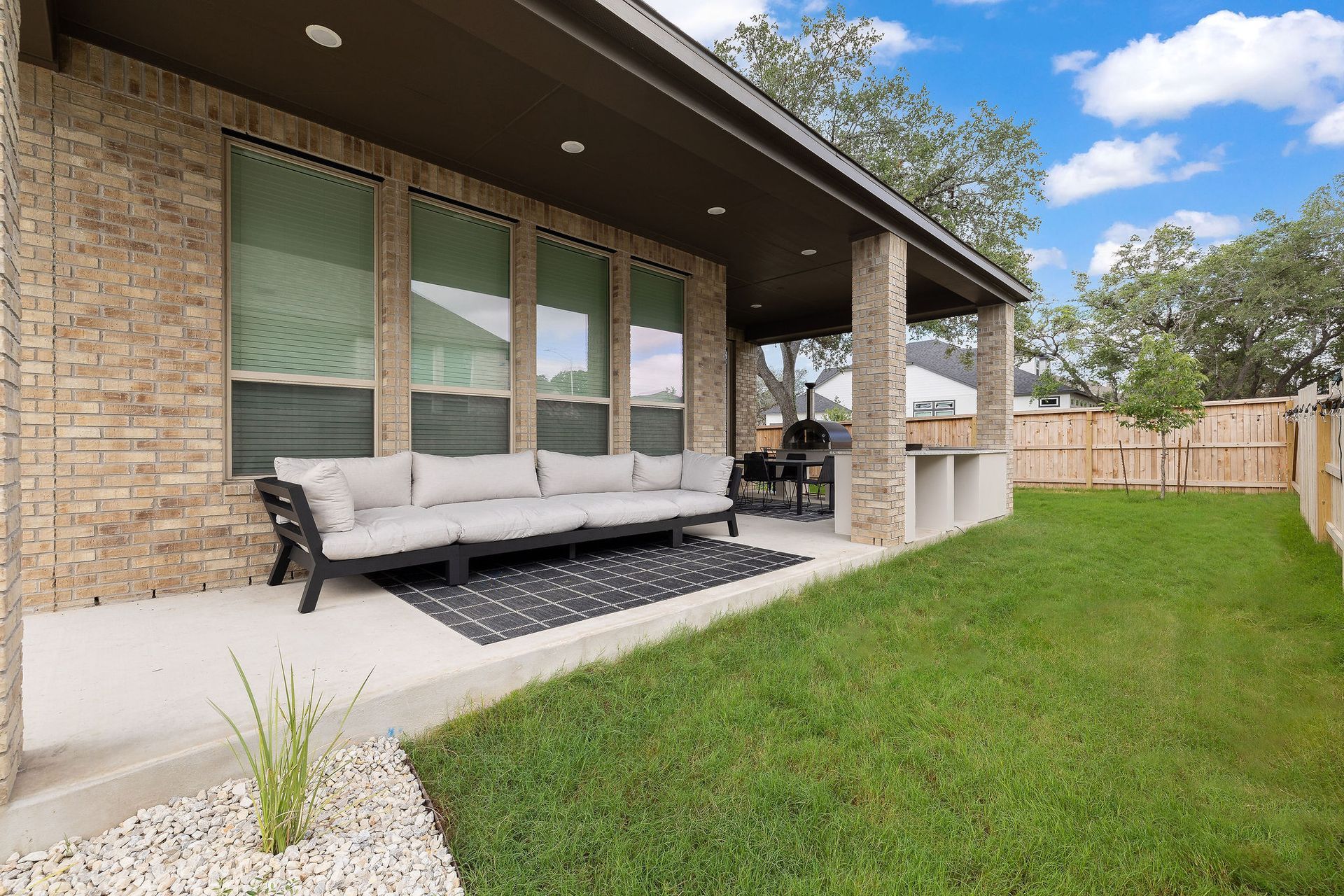 Covered patio with outdoor sofa, grill, and green lawn beside a brick house